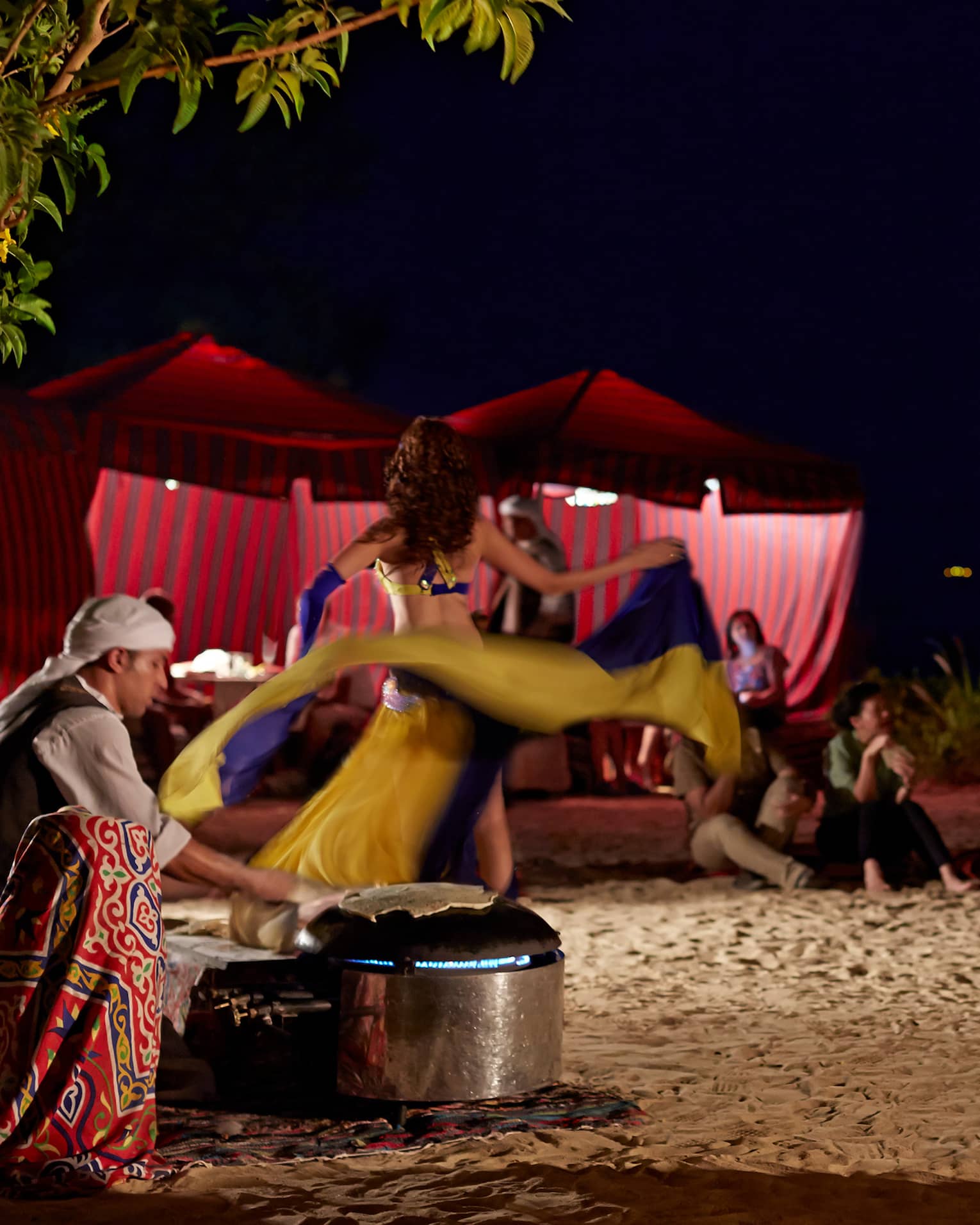 At night, a dancer performs outdoors for guests, swirling their skirt to music played by three smiling, seated musicians.