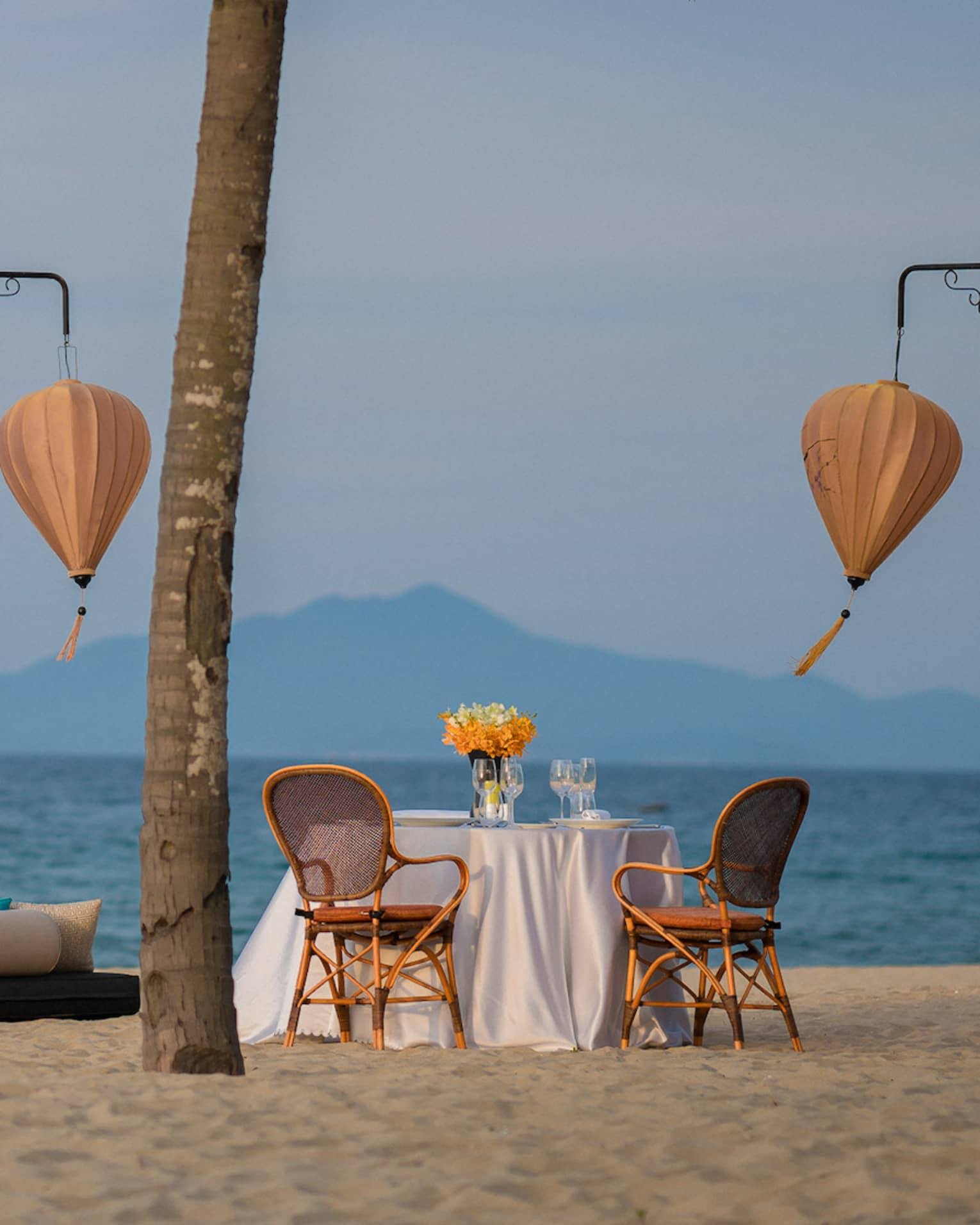 Couple walks on sandy beach by dining table between two palm trees, paper lanterns