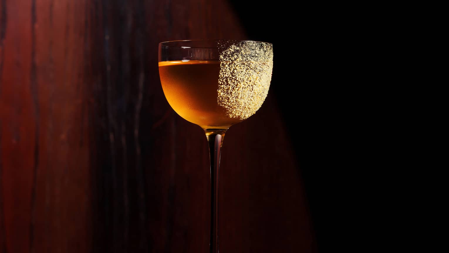 A tall stemmed red wine glass holds a golden-colored cocktail with gold dust sprinkled on the right side of the glass