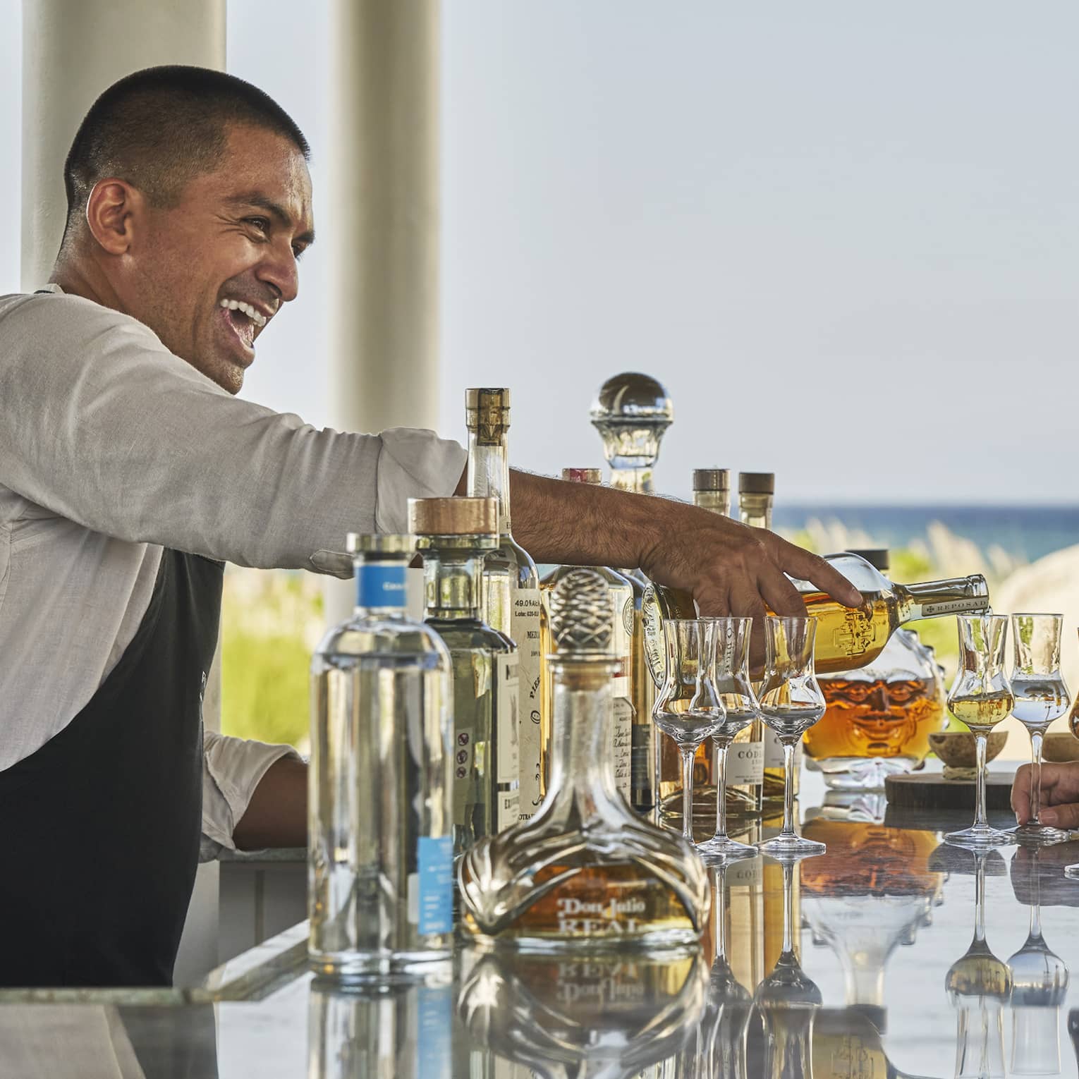 A male bartender serving drinks to a man and woman at a bar next to a beach.