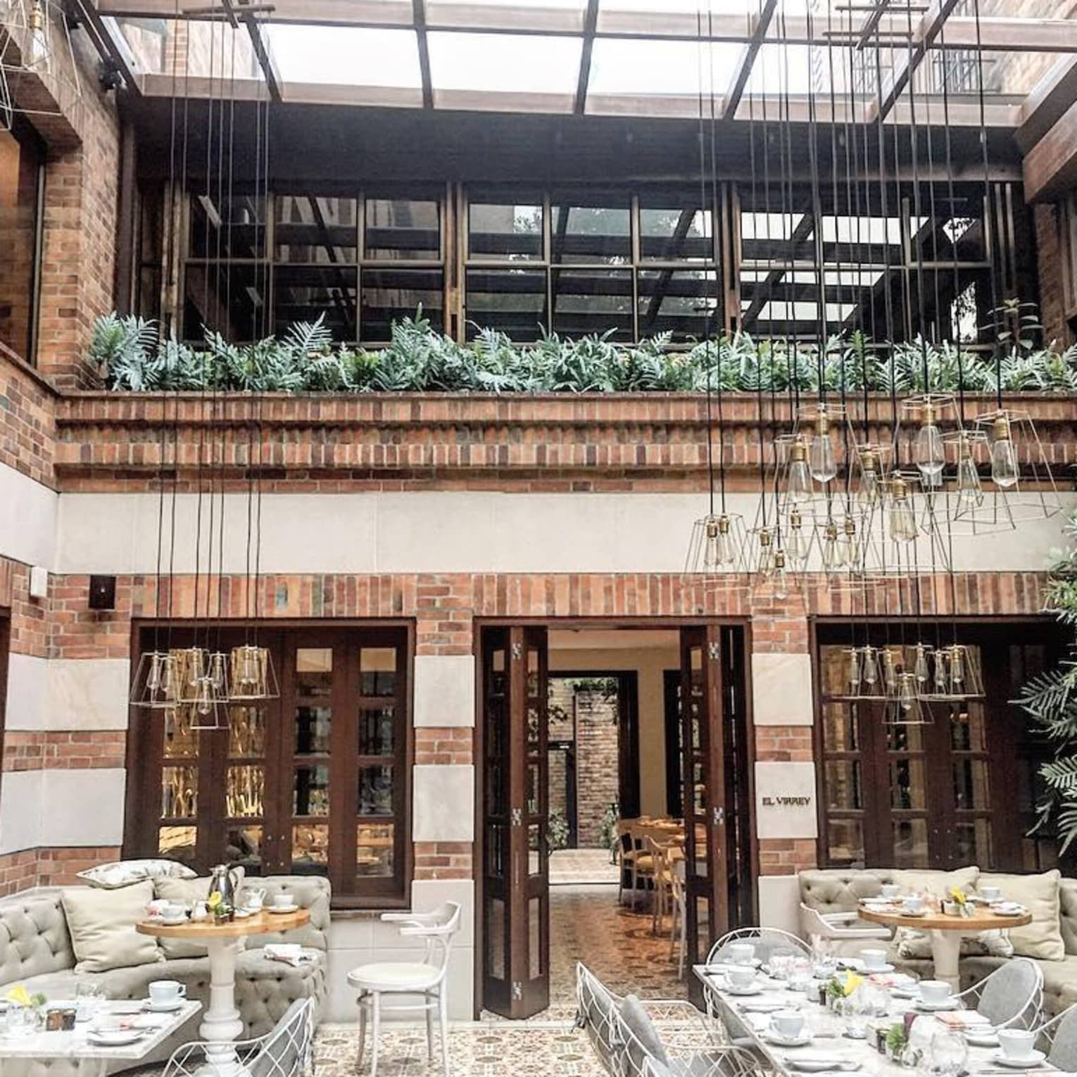 Dining tables and chairs under sunny skylight, brick indoor balcony lined with tropical plants