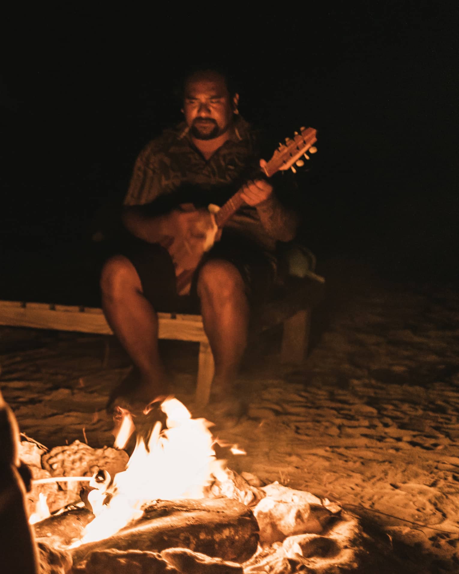 Group of people sitting around a campfire at night, with one person playing a guitar in the background