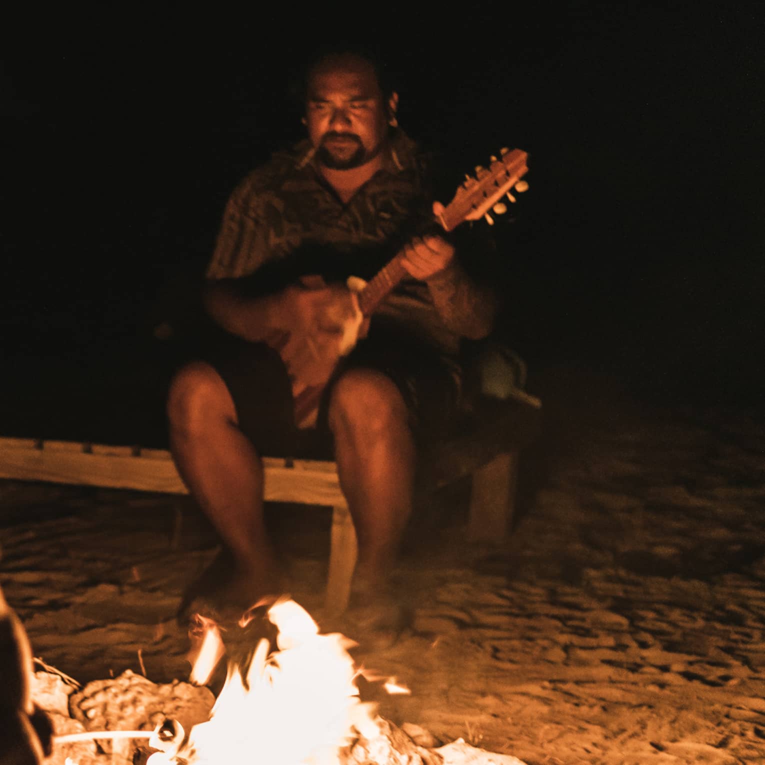 Group of people sitting around a campfire at night, with one person playing a guitar in the background