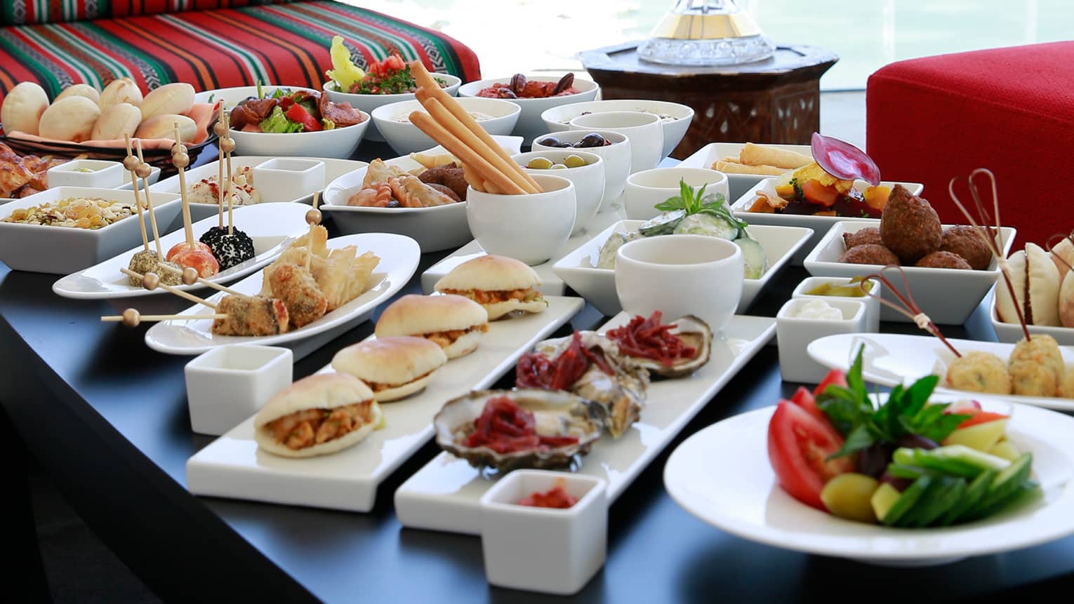 Table with more than a dozen white dishes with mezze appetizers and snacks, striped sofa and red bench