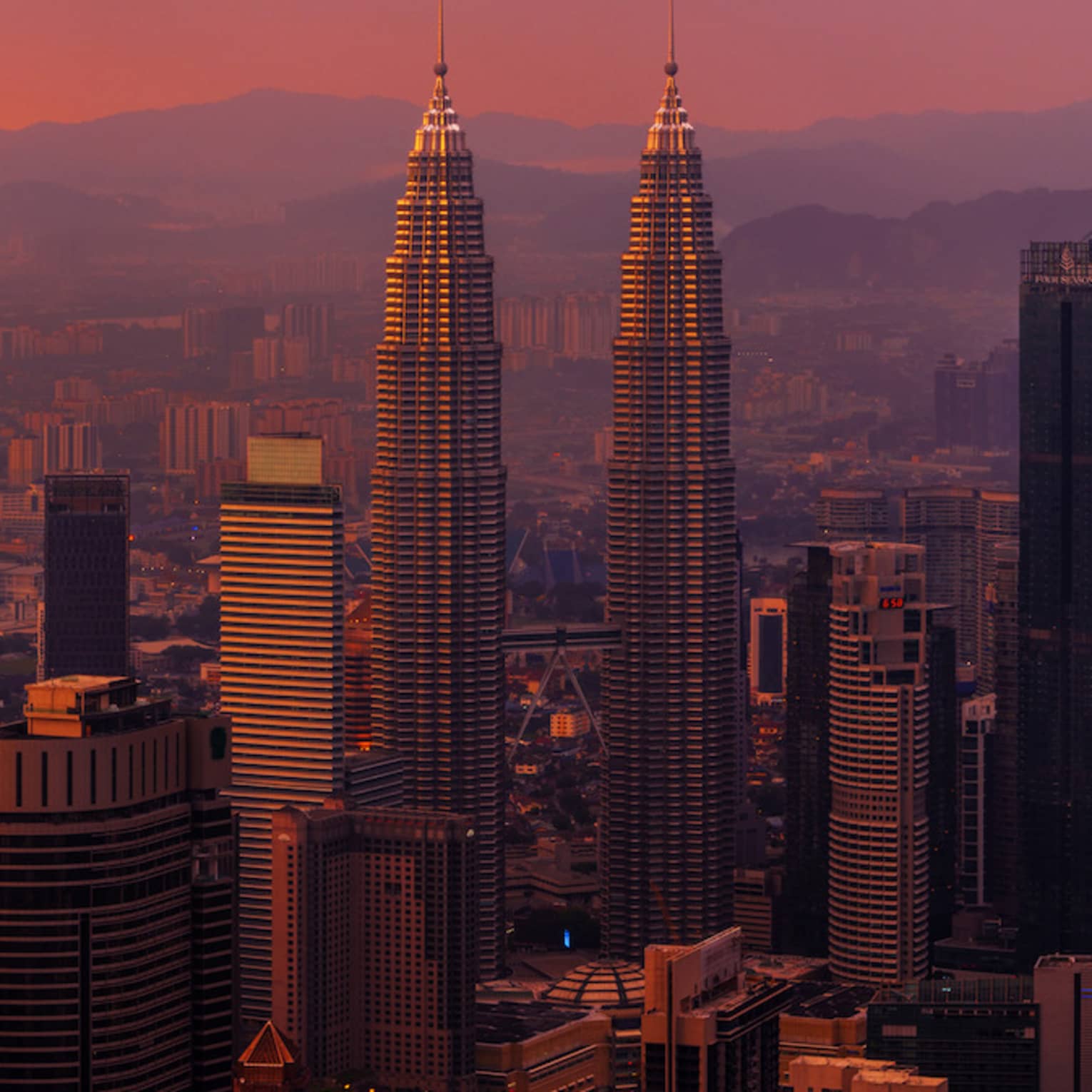 Majestic towers mark the city skyline. Reds and yellows blend in a magnificent sunset as mountains loom in the horizon.