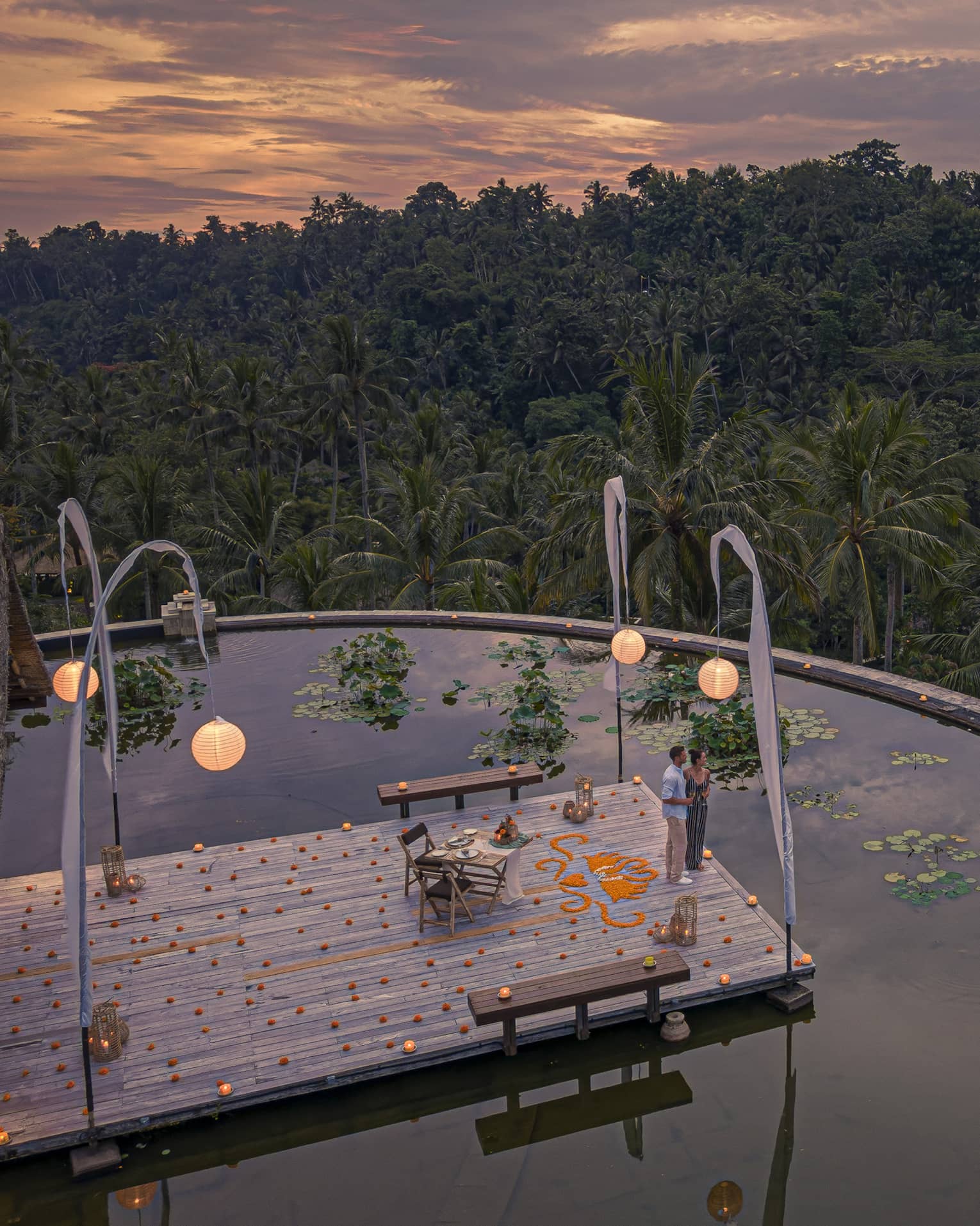 Couple stands on dock at the infinity edge lotus pond overlooking dense forest at dusk