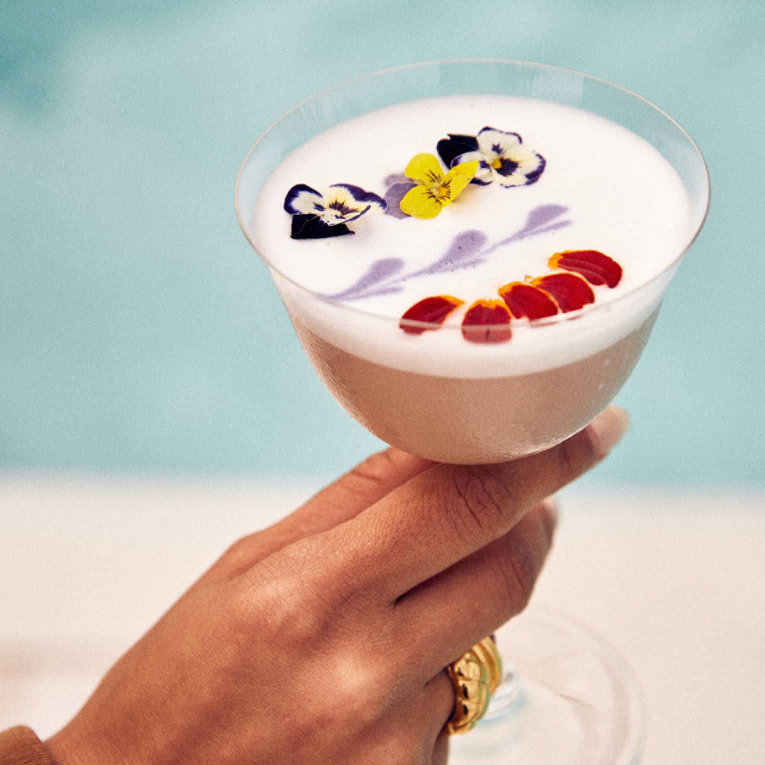 An egg white cocktail decorated with edible yellow, purple and white flowers, along with red petals and purple art on top.