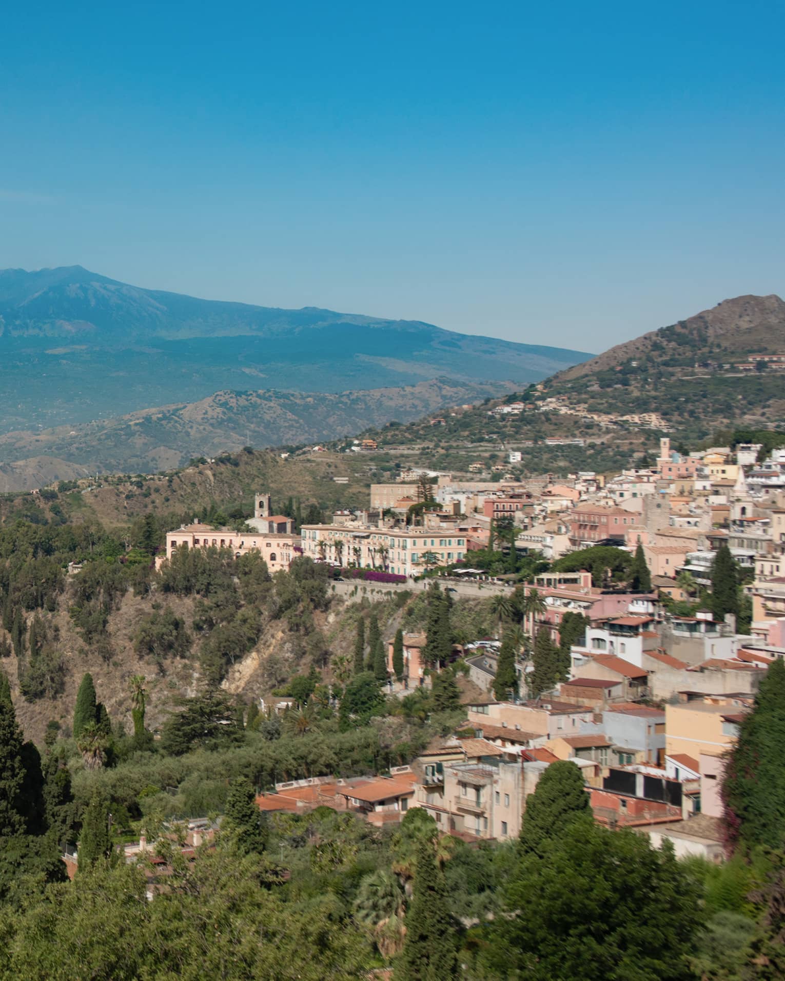Panoramic view of a coastal town nestled in the hills, overlooking the sea, with mountains in the background and greenery in the foreground.