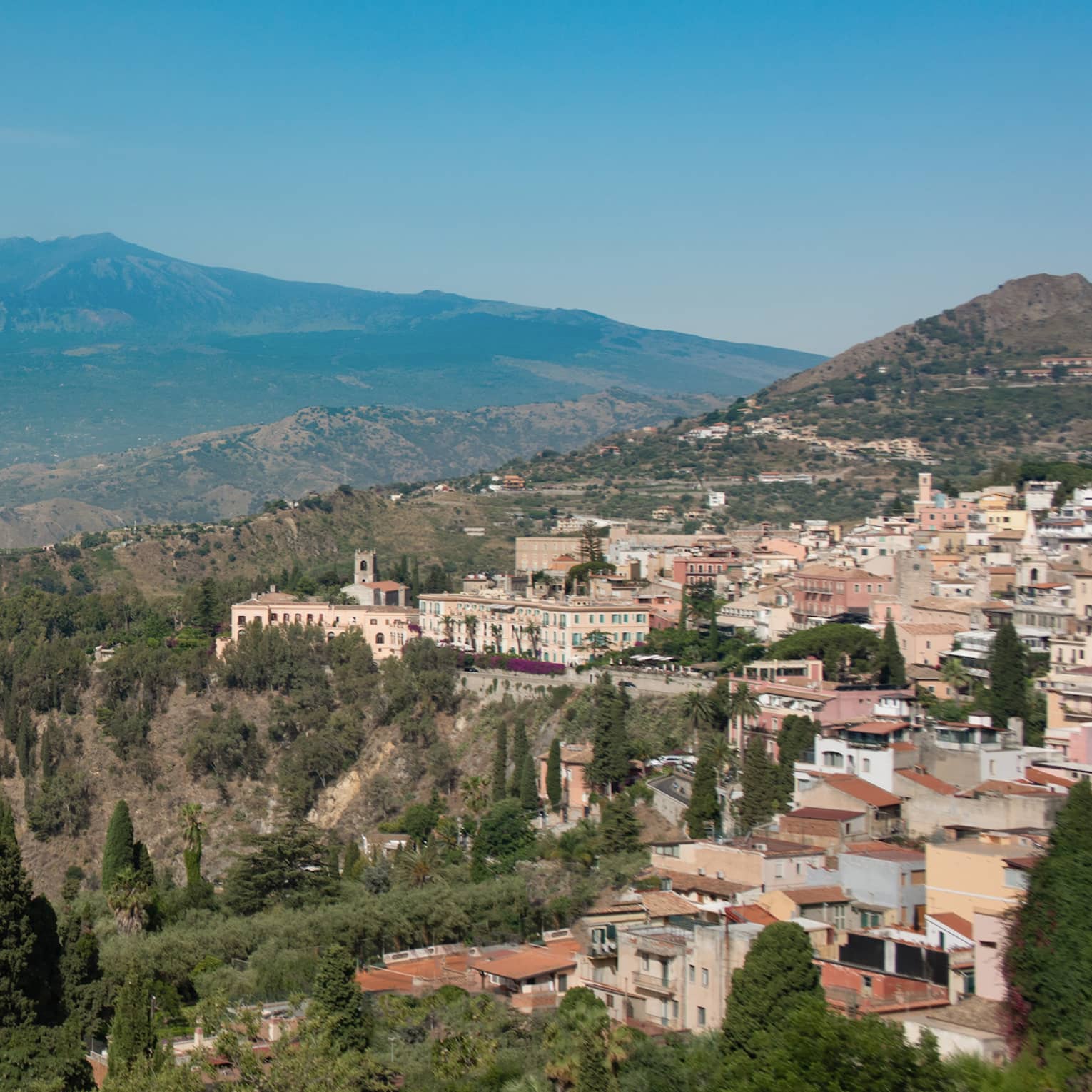 Panoramic view of a coastal town nestled in the hills, overlooking the sea, with mountains in the background and greenery in the foreground.