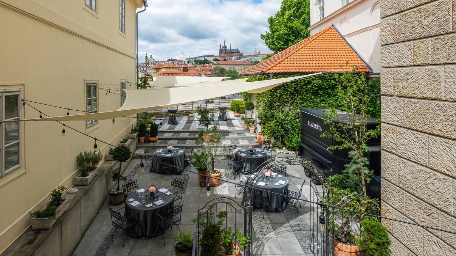 Piazzetta rooftop restaurant with black round iron tables, topiaries, Prague Castle view