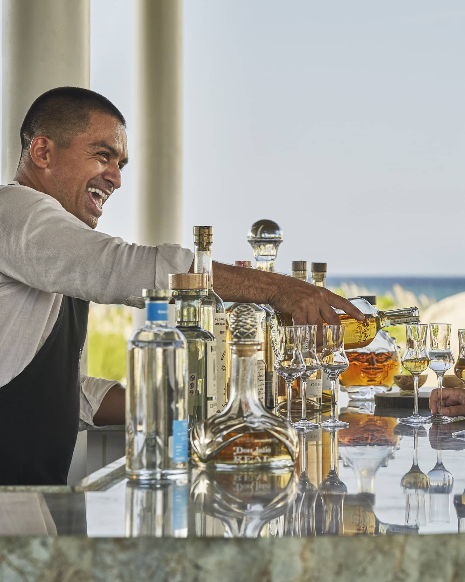 A male bartender serving drinks to a man and woman at a bar next to a beach.