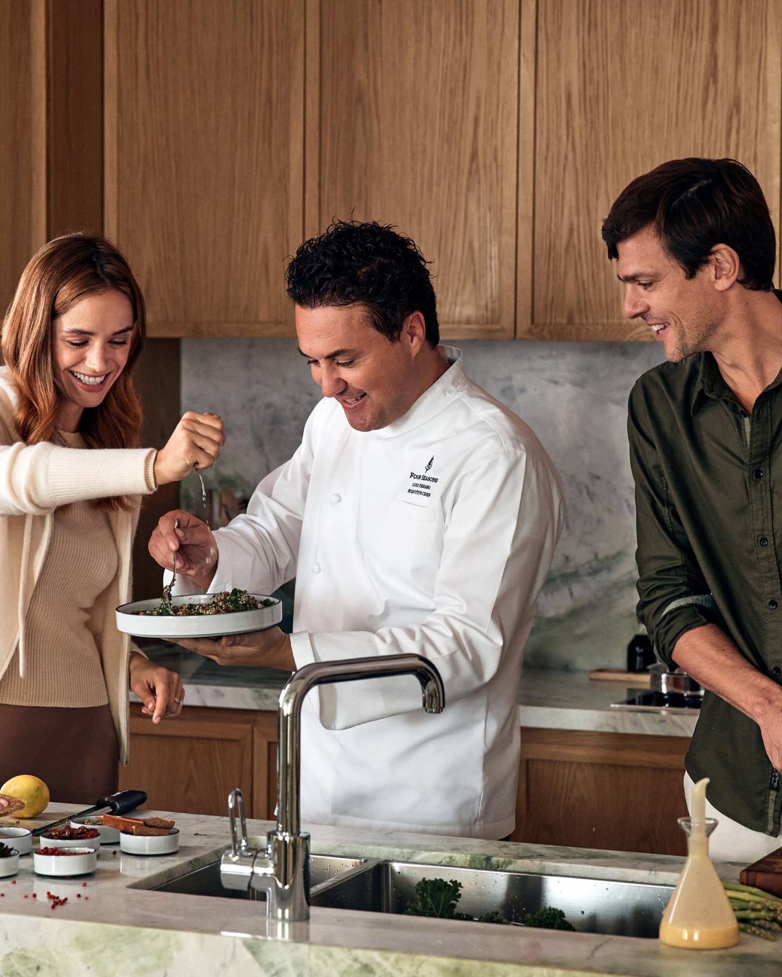 A chef holds a plate of tabbouleh as a guest squeezes a lemon overtop and another guest chops vegetables on a marble counter.