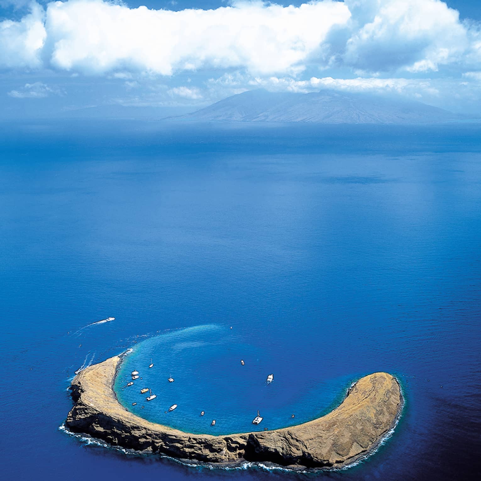 Aerial view of Molokini Crater, a crescent-shaped volcanic islet surrounded by clear blue ocean waters, with boats anchored nearby