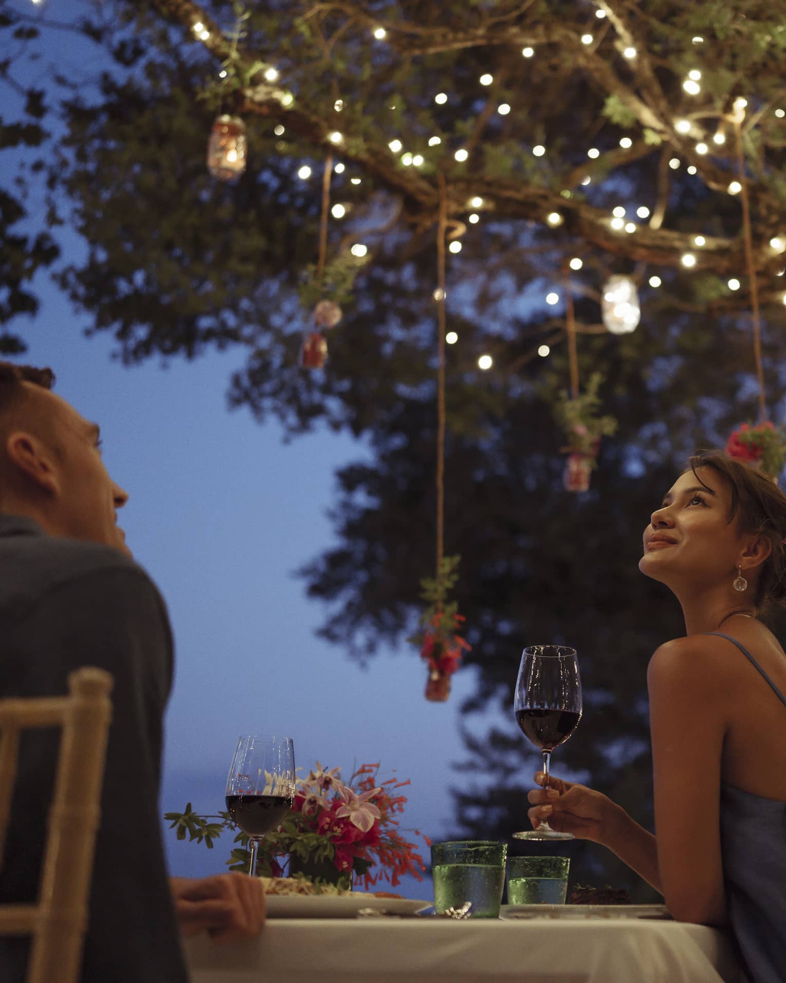A smiling couple having a romantic dinner under trees covered in string lights and jars of flowers hanging from the branches.