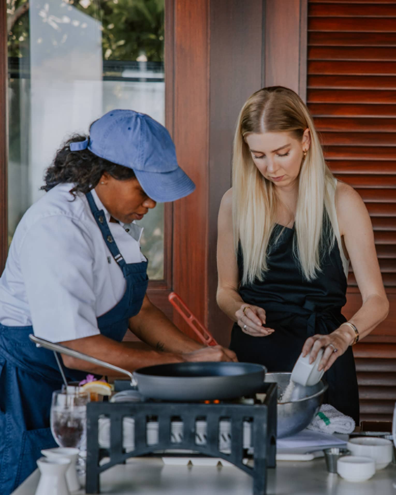 A cooking class is underway as three people sporting aprons stand over a table strewn with wine glasses and cookware.