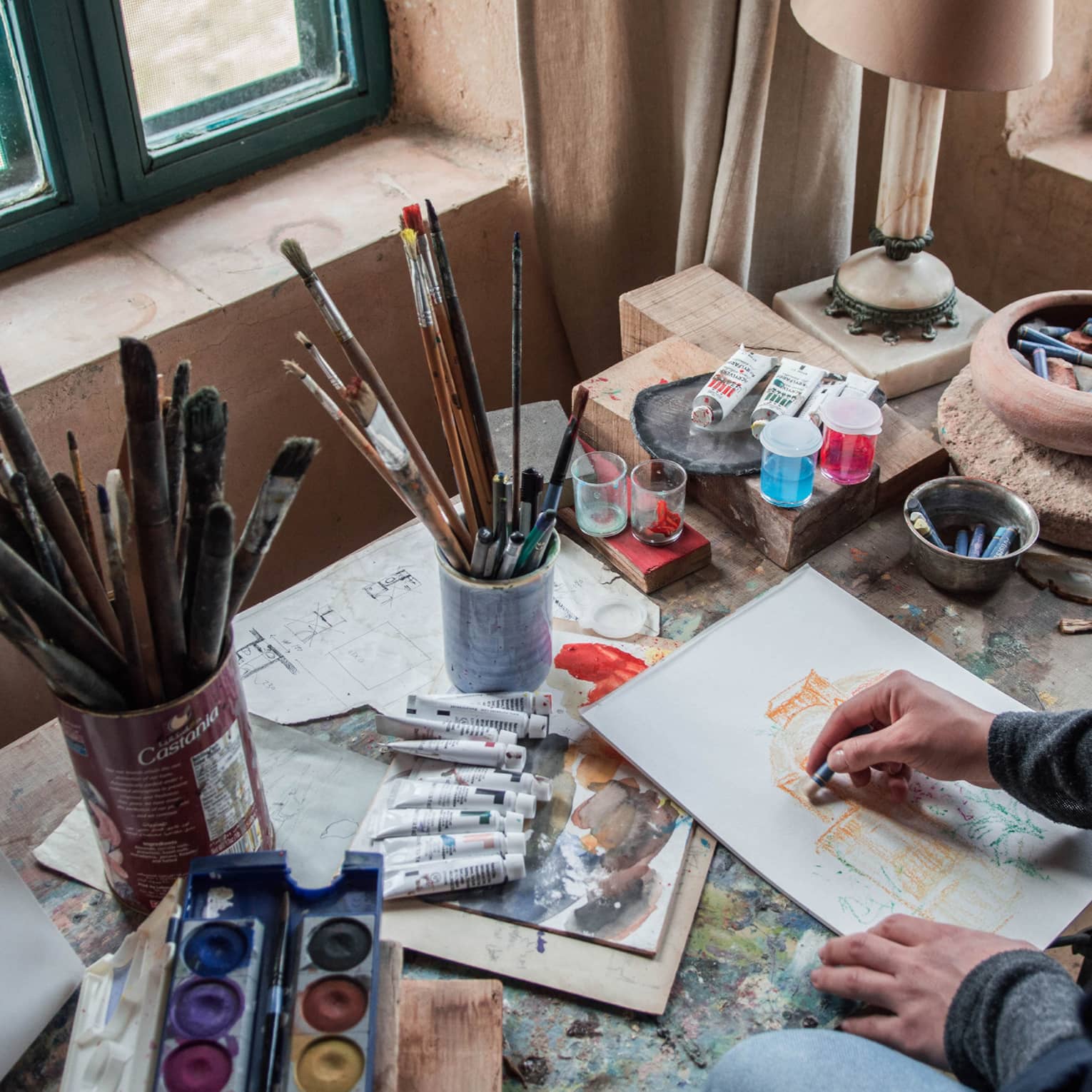 A person with dark curly hair in front of an artists workstation with paintbrushes and acrylic paints.