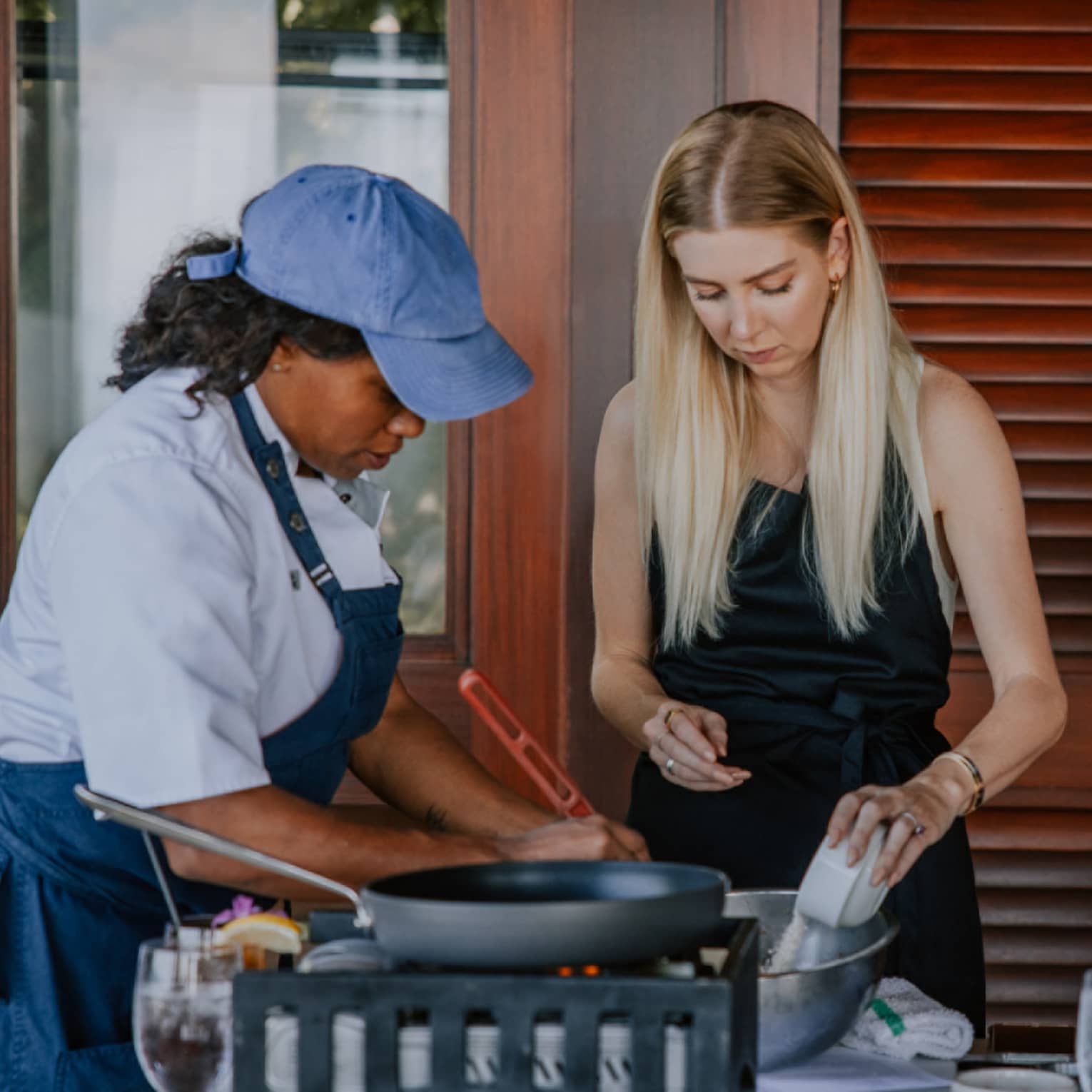 A cooking class is underway as three people sporting aprons stand over a table strewn with wine glasses and cookware.