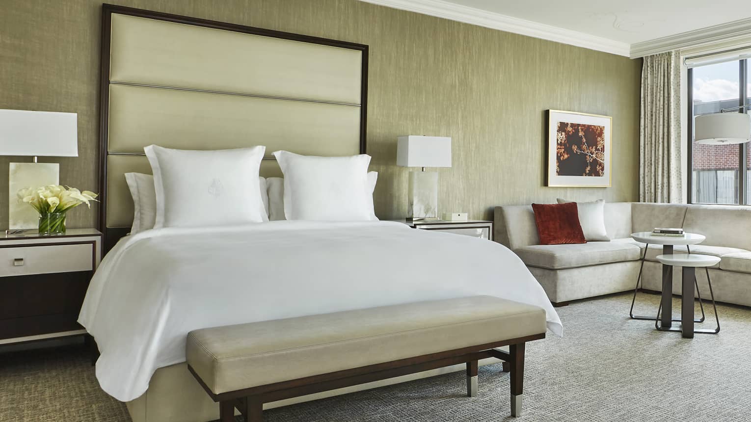Grand Premier Room bed with white bench, tall padded headboard, L-shaped sofa near sunny picture window