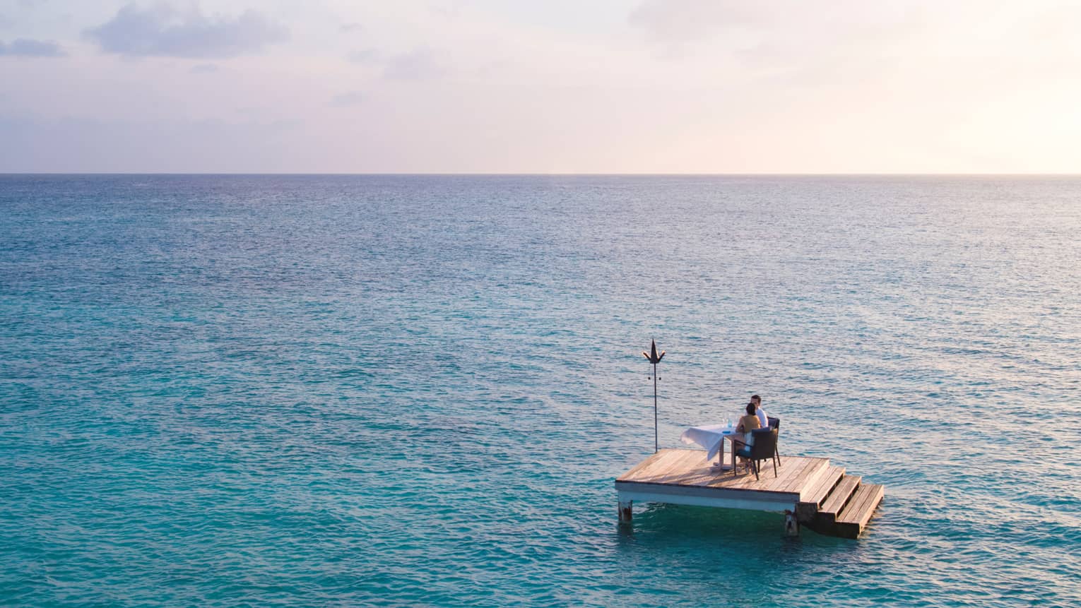 Man, woman dine at private table on small floating wood platform in middle of blue lagoon