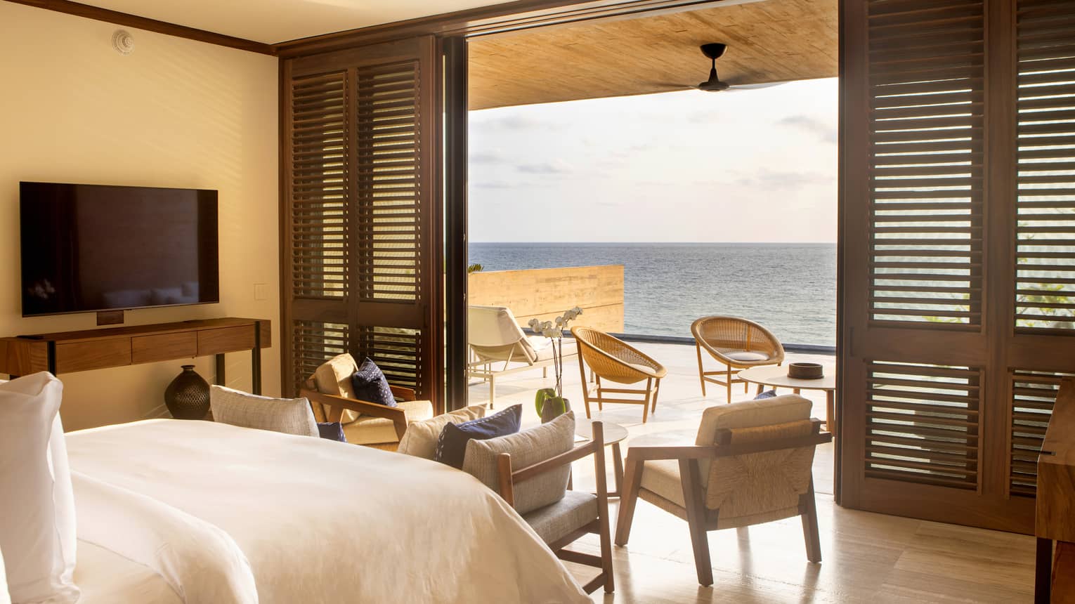Bedroom with bed and white linens, armchair and flat screen TV, sliding doors opening onto terrace with ocean views