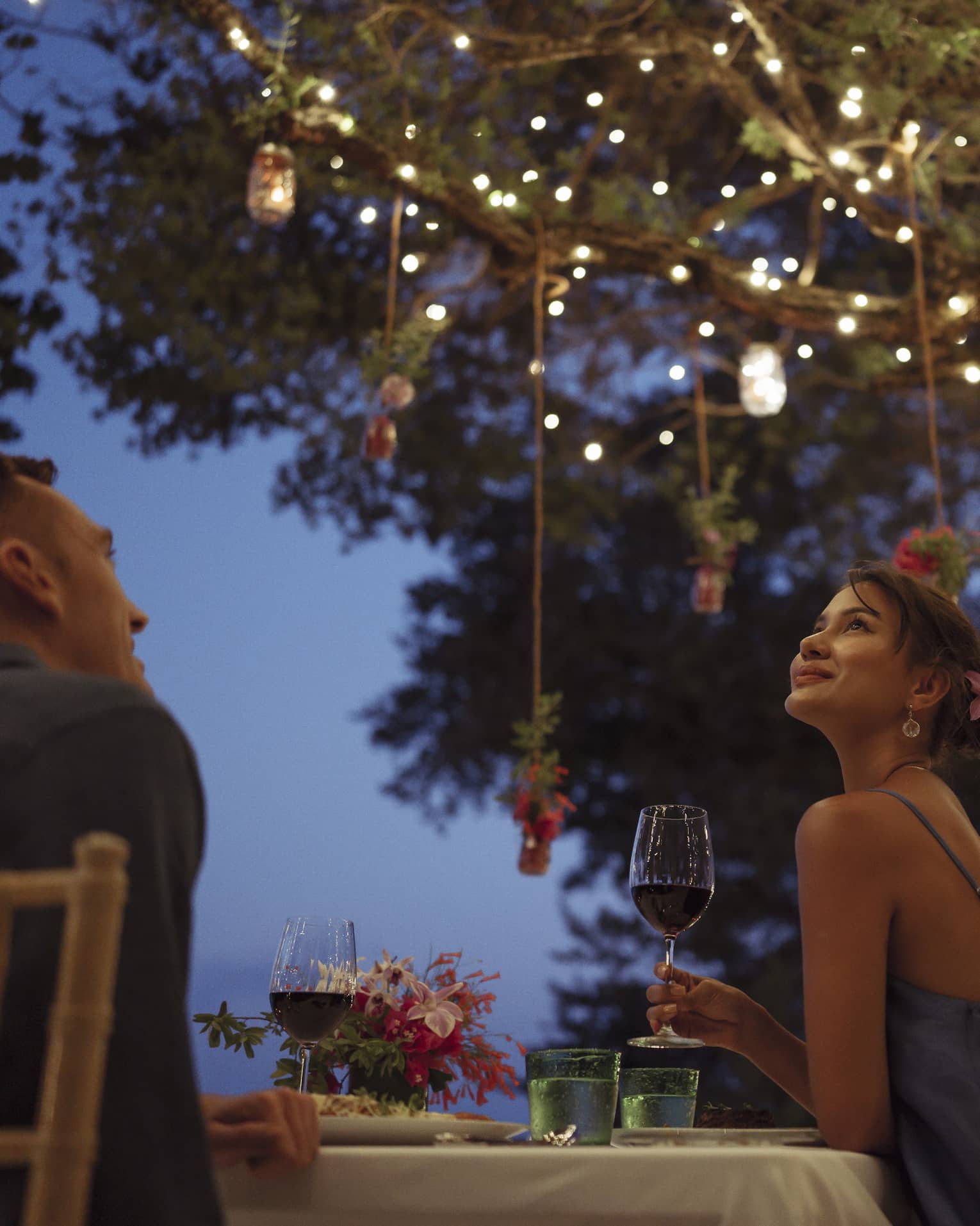 A smiling couple having a romantic dinner under trees covered in string lights and jars of flowers hanging from the branches.