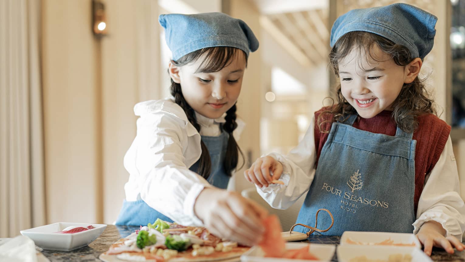 Two young girls in matching denim Four Seasons aprons and bandanas add toppings to their pizzas