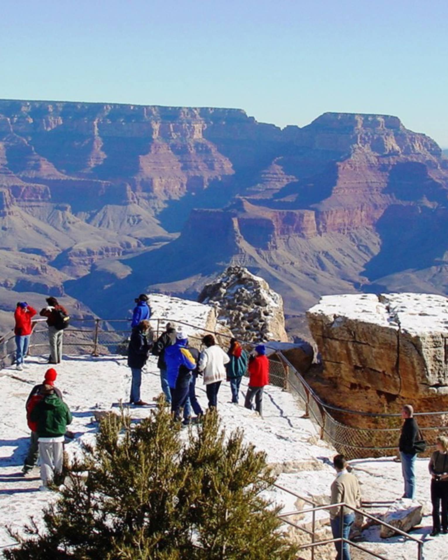 Tourists on a fenced platform, some taking photos, dwarfed by the the majestic Grand Canyon backdrop beneath a clear sky.