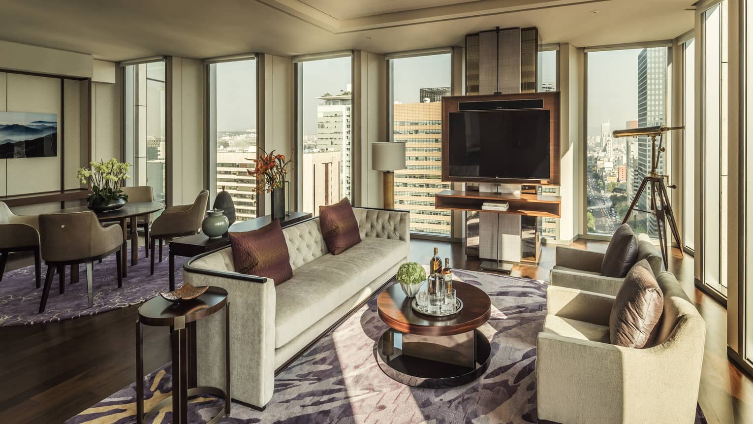 Ambassador One-Bedroom Suite long sofa, brown silk pillows, two armchairs around table with liquor, corner windows and telescope