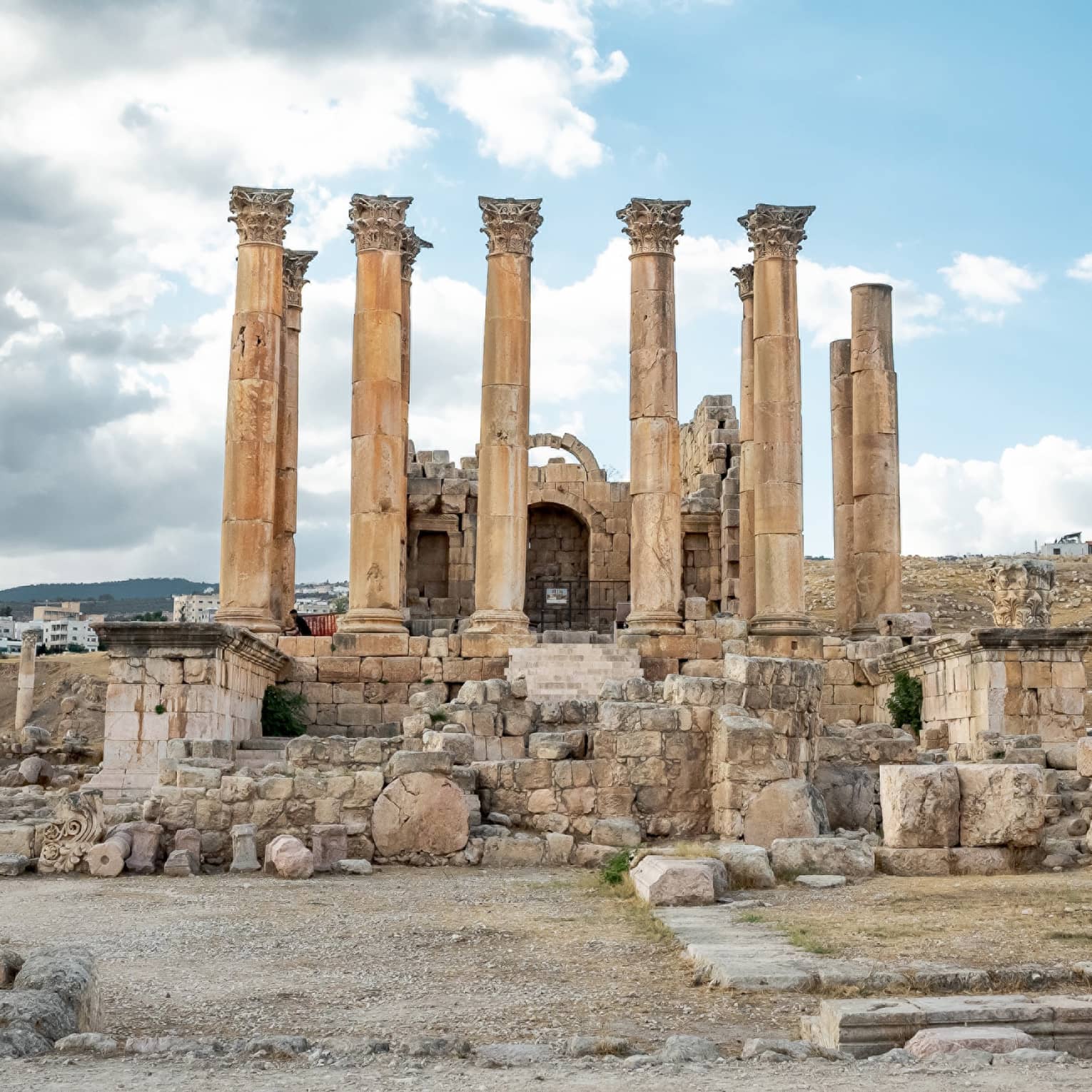 Amongst crumbling ruins, ten ancient Corinthian columns stretch skyward under a cluster of grey and white clouds.