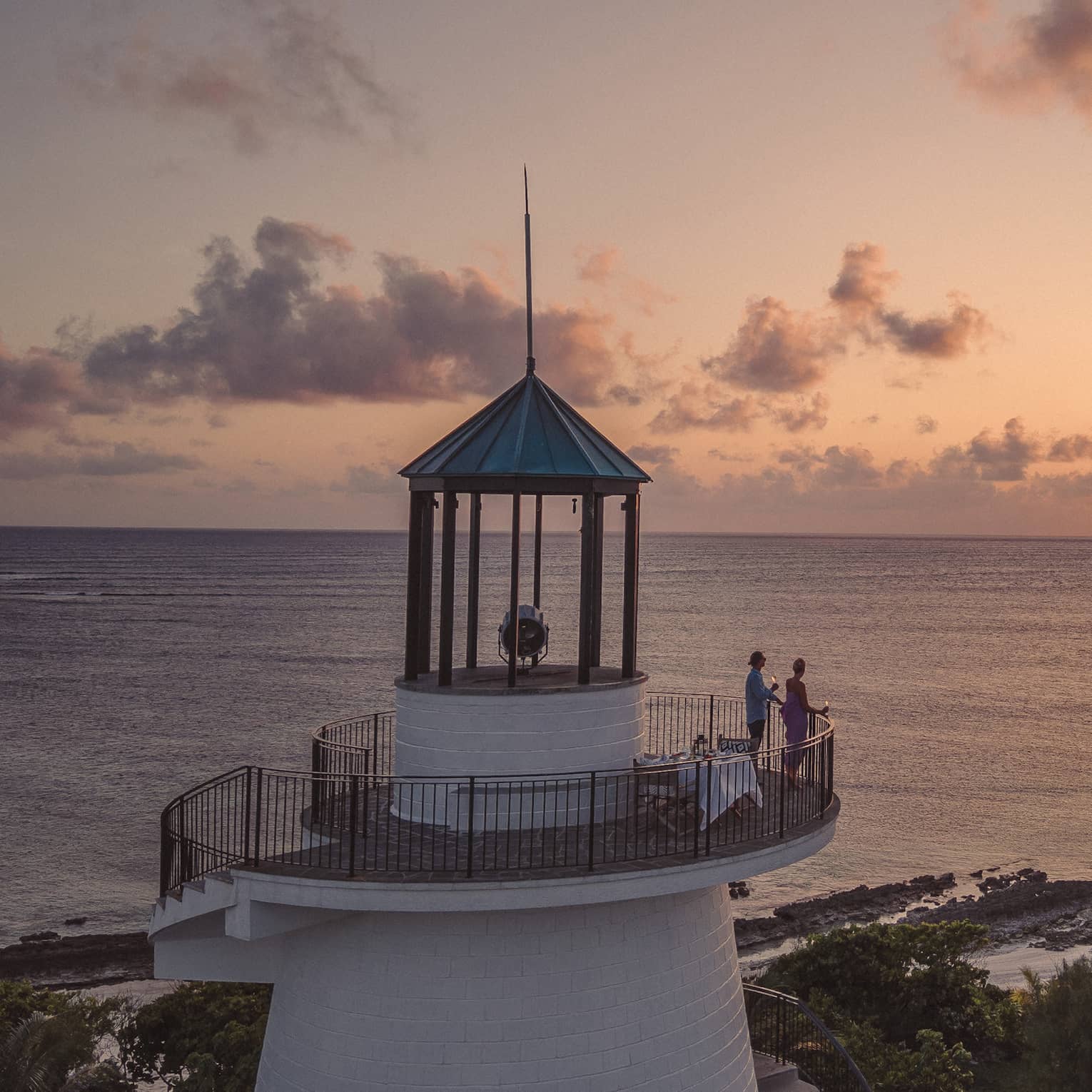 Long view of two people on a beach lighthouse balcony, enjoying the sunset over the ocean, distant silhouette of birds.