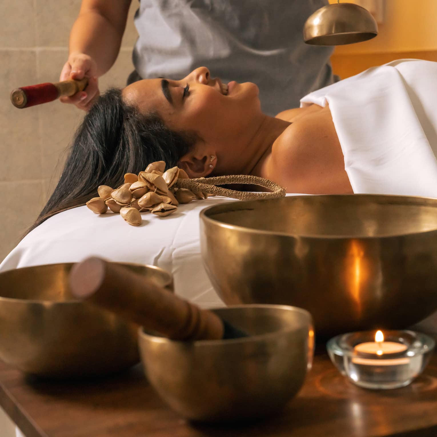 Three Tibetan sound bowls sit on a table next to a person lying beneath a white sheet while another person rings a smaller bowl over them