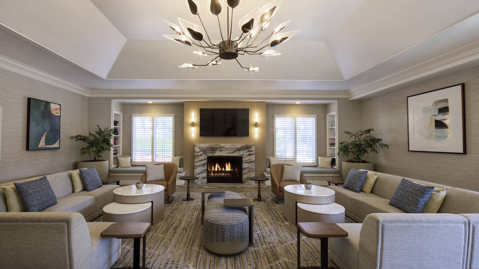 A spacious and inviting room with neutral decor and an artistic chandelier features couches with pillows, low tables, art on the walls, a fireplace, large potted plants, a flat-screen TV and windows admitting natural light.