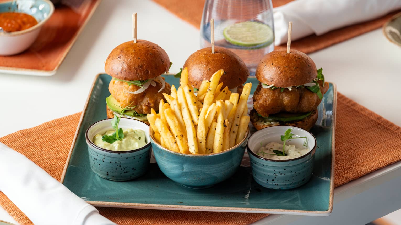 Three crispy fish sliders served with a bowl of shoestring fries and two dipping sauces on a blue plate