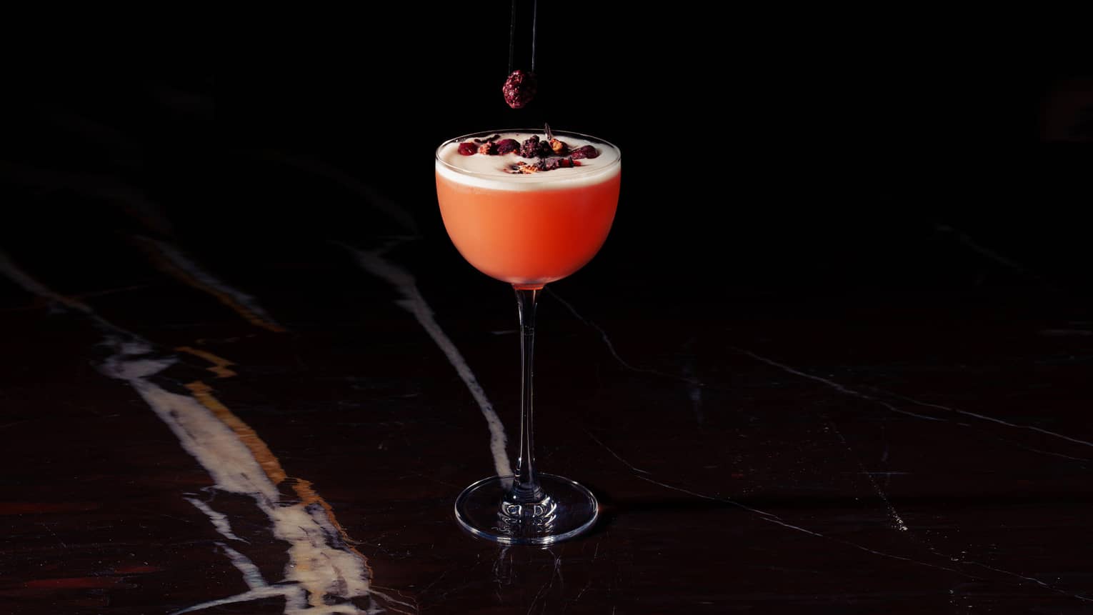 A hand reaches out of the darkness to drop dried cranberries onto a red-colored cocktail in a tall stemmed glass