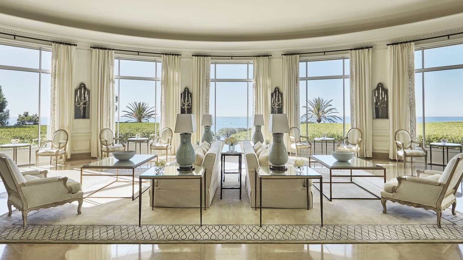 Rotonde seating area with white sofas, chairs, lamps and bright windows overlooking lawns, sea