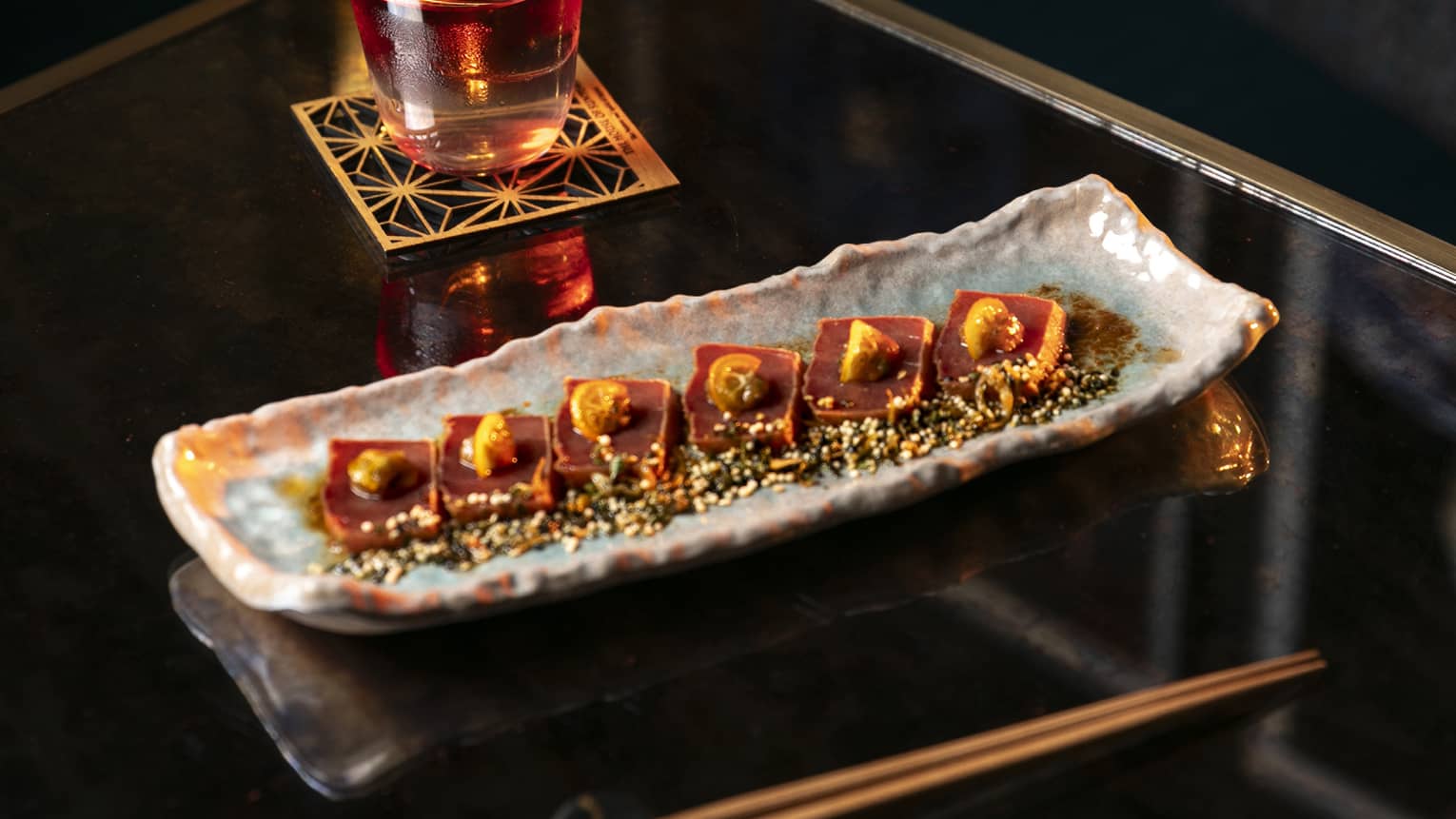 An artistic rectangular glazed plate holds a row of food items and is set on a table next to a drink and a pair of chopsticks.