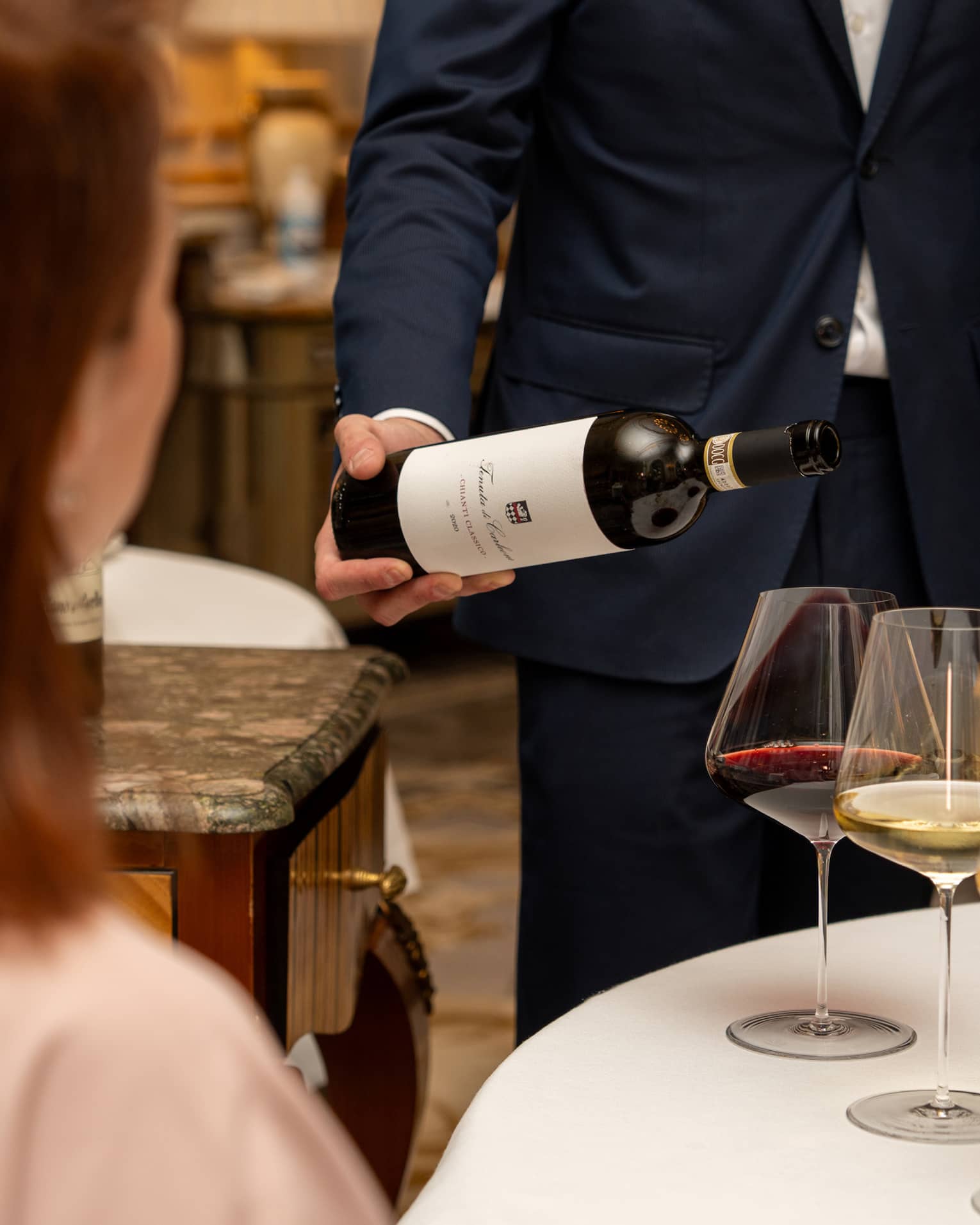 A guest sits at a table with three glasses of wine as a smiling sommelier tilts a bottle of red wine over one of the glasses.