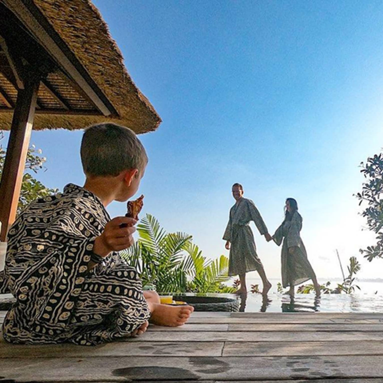 Family wearing robes, young child sits cross legged on patio as parents hold hands in background