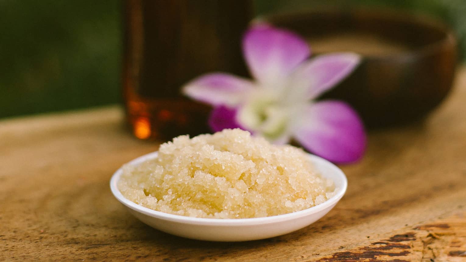 Bowl with salt scrub next to purple tropical flower and glass oil bottle on rustic table