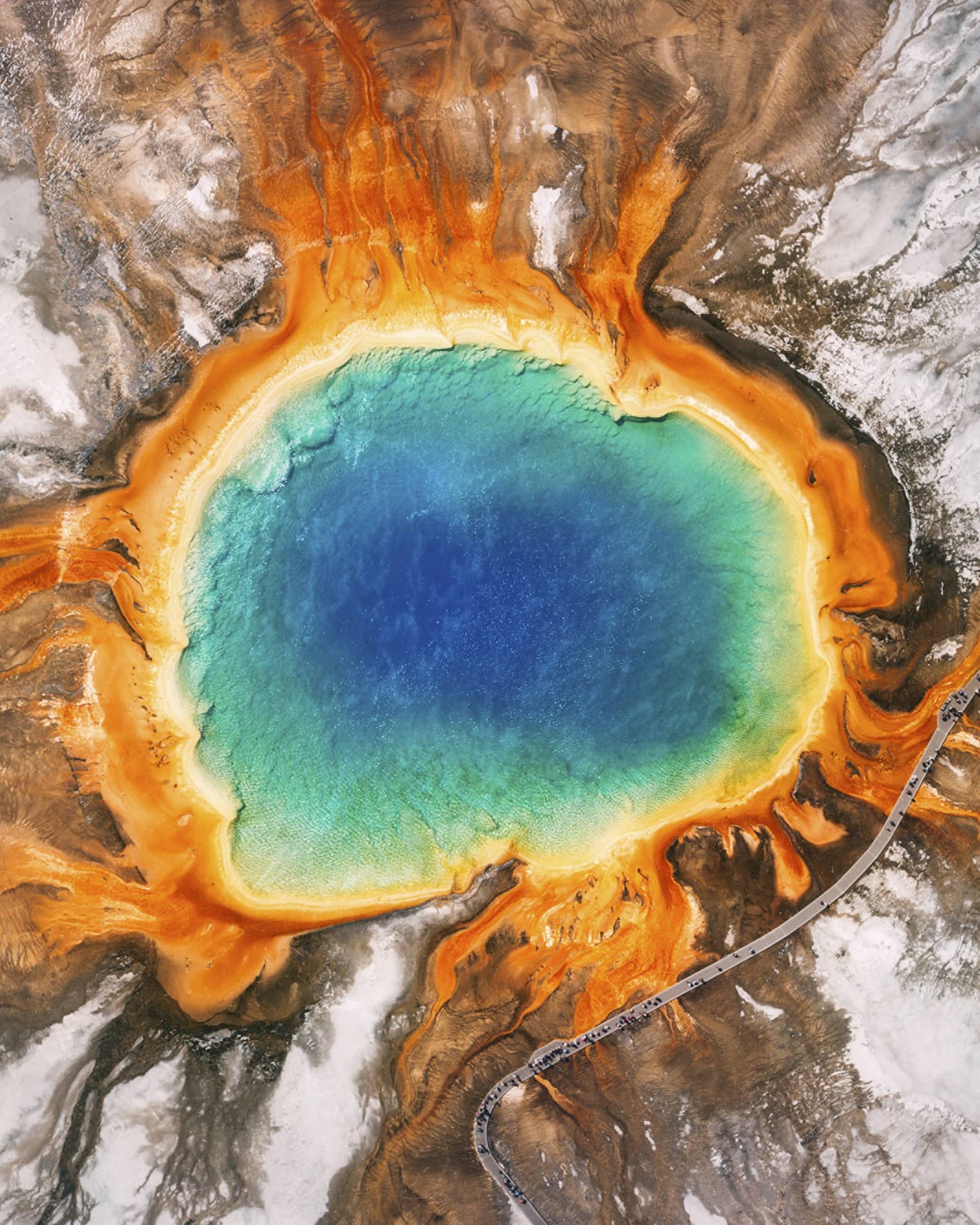 Aerial view of the Grand Prismatic Spring in Yellowstone, featuring vibrant blue, green, yellow and orange concentric rings, surrounded by rocky terrain