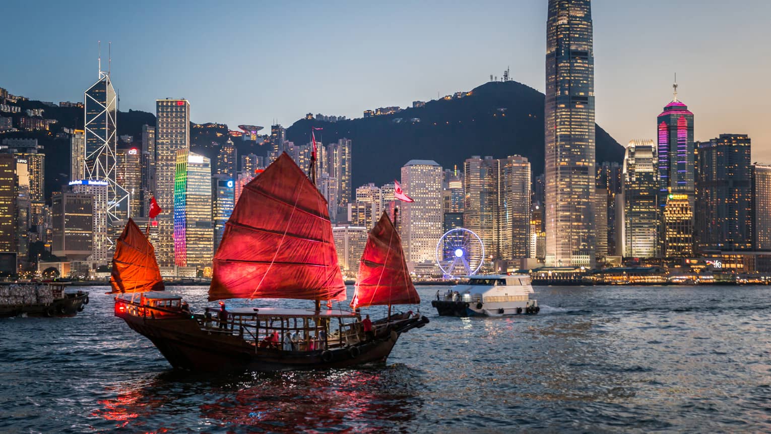 A traditional Hong Kong junk boat with red sails gliding through Victoria Harbour at dusk, with the city’s illuminated skyline in the background