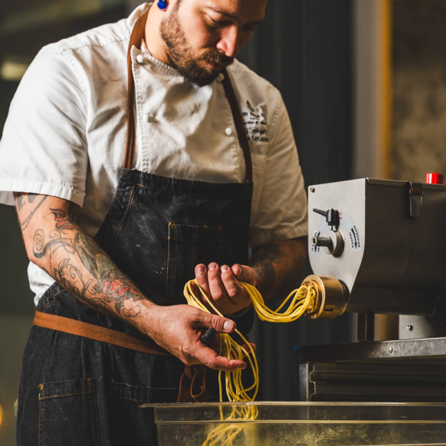 A chef catches fresh long pasta noodles as they emerge from an industrial pasta-making machine in a warmly lit kitchen.