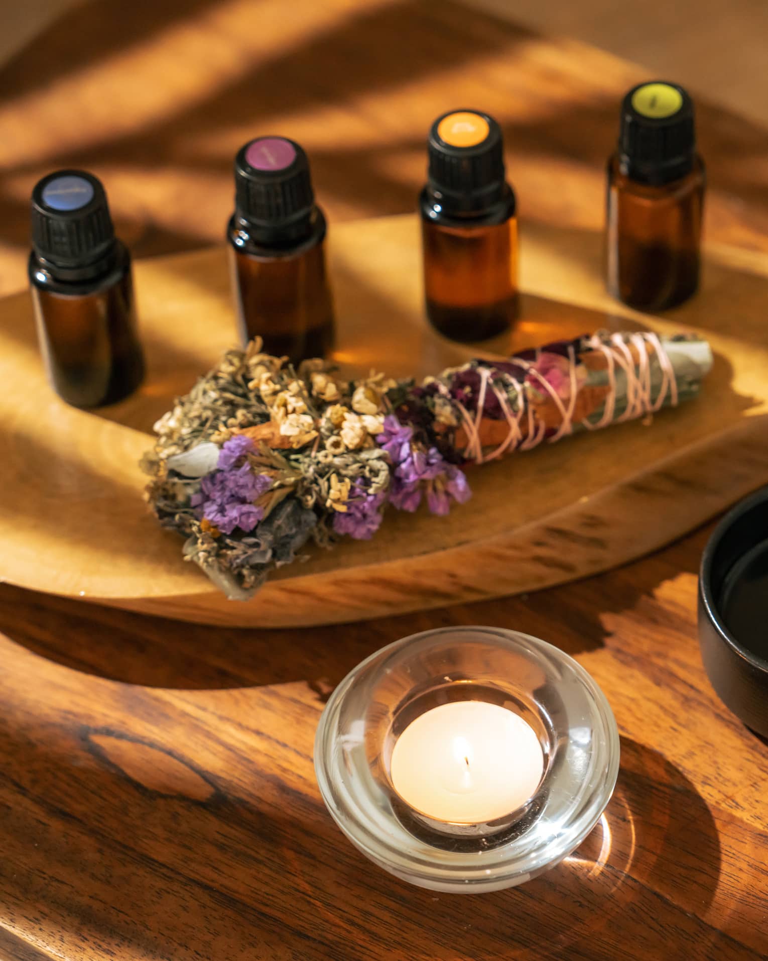 Four small bottles of essential oils and a sage-and-floral bundle stick lie on a wooden platter next to a hand pouring liquid into a small black bowl next to a votive candle
