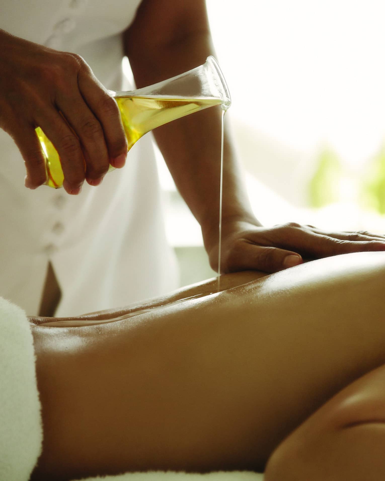 Spa staff pours massage oil over woman's bare back as she lays on treatment bed