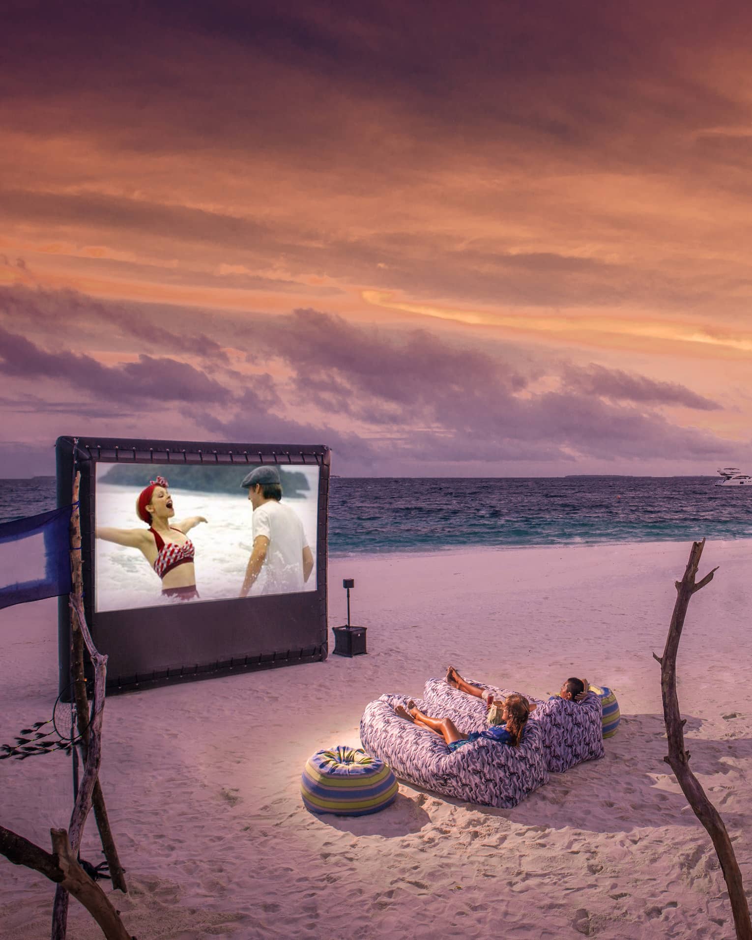 ,Two people in beanbag chairs enjoy a movie night on the beach at dusk, popcorn stand on sand behind them