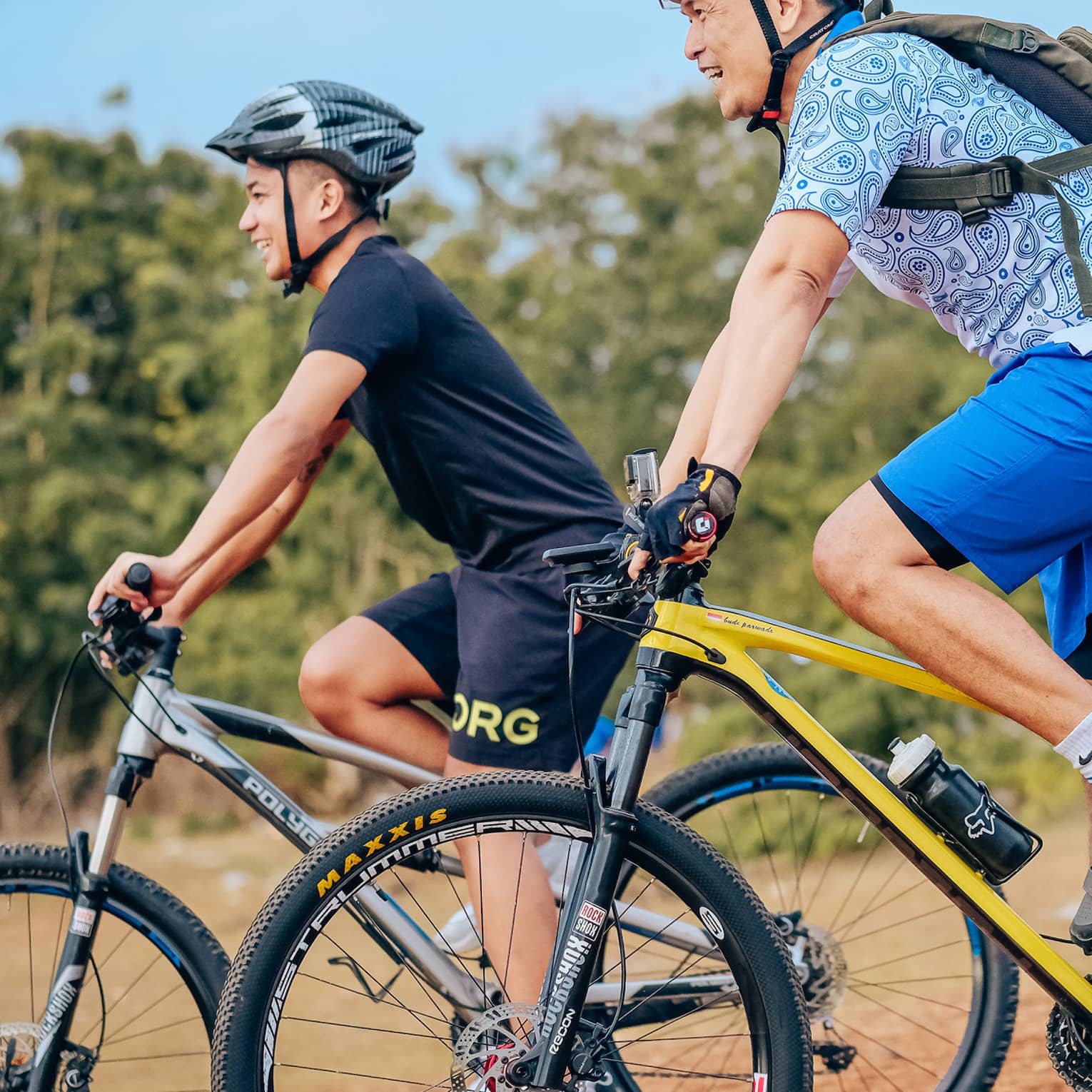 Side view of two smiling mountain bikers in helmets as they coast down a gentle slope on a dirt path alongside trees.