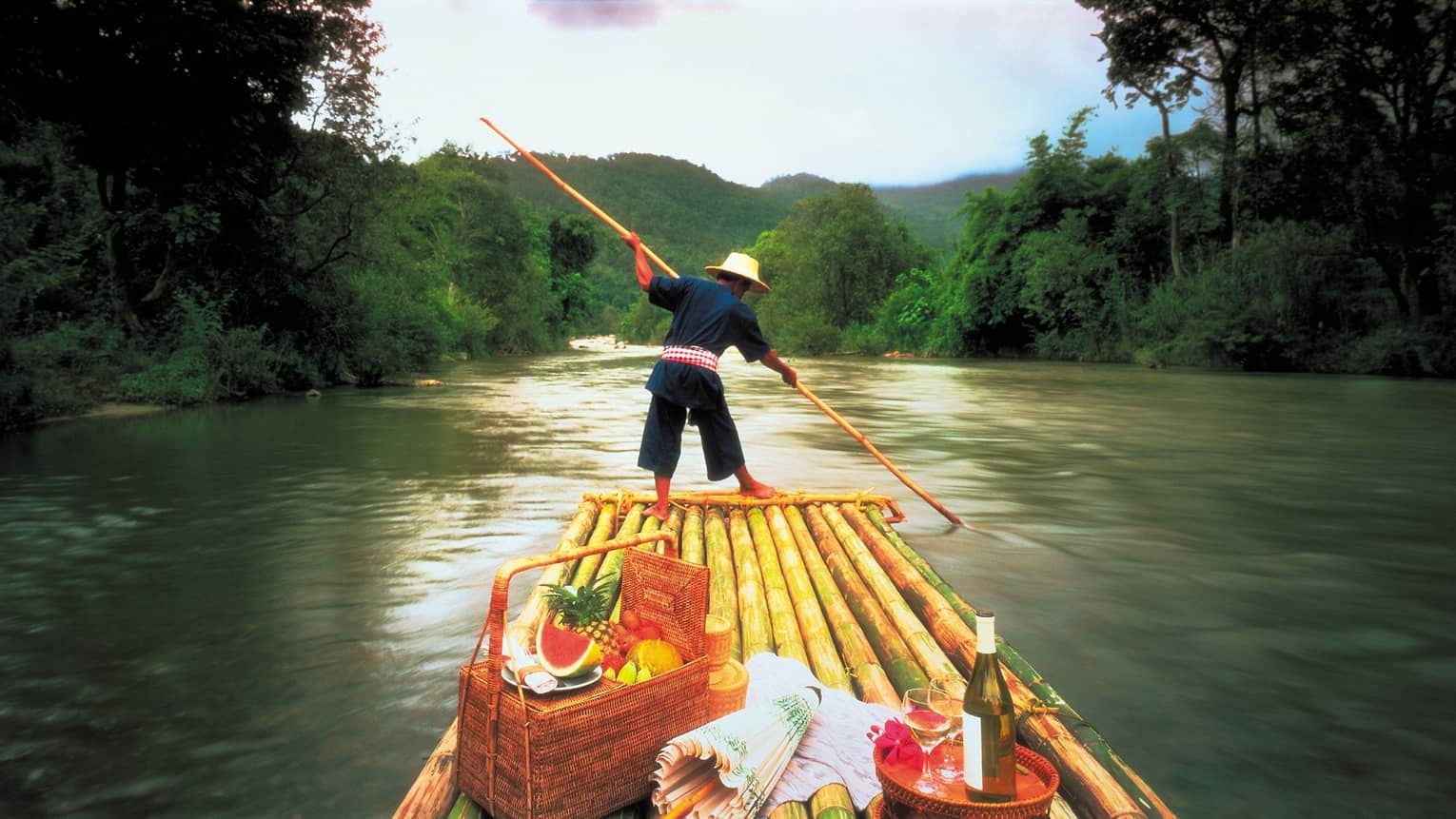 Man steers boat down Mae Tang River in front of picnic baskets, fruit and wine
