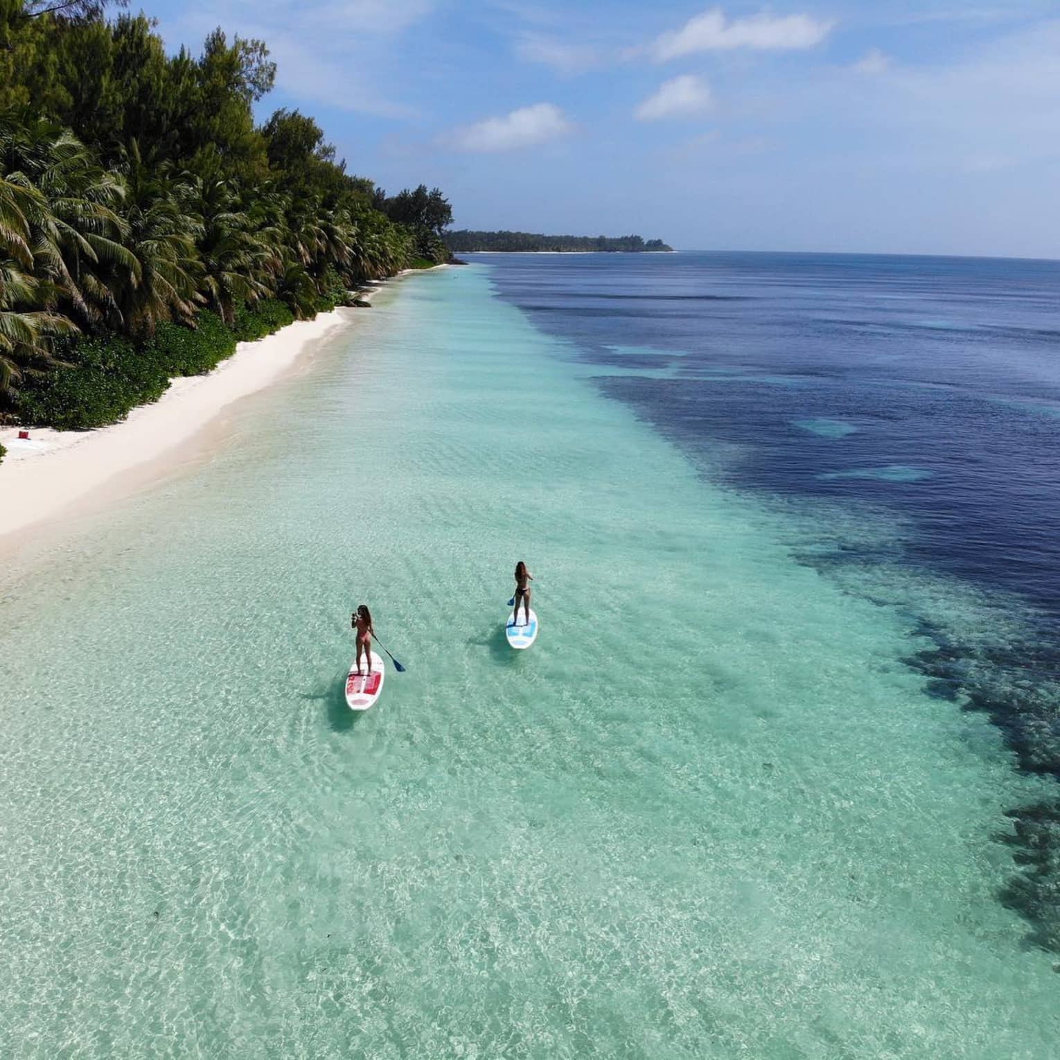 Two women on paddle boards float through crystal clear water next to the beach