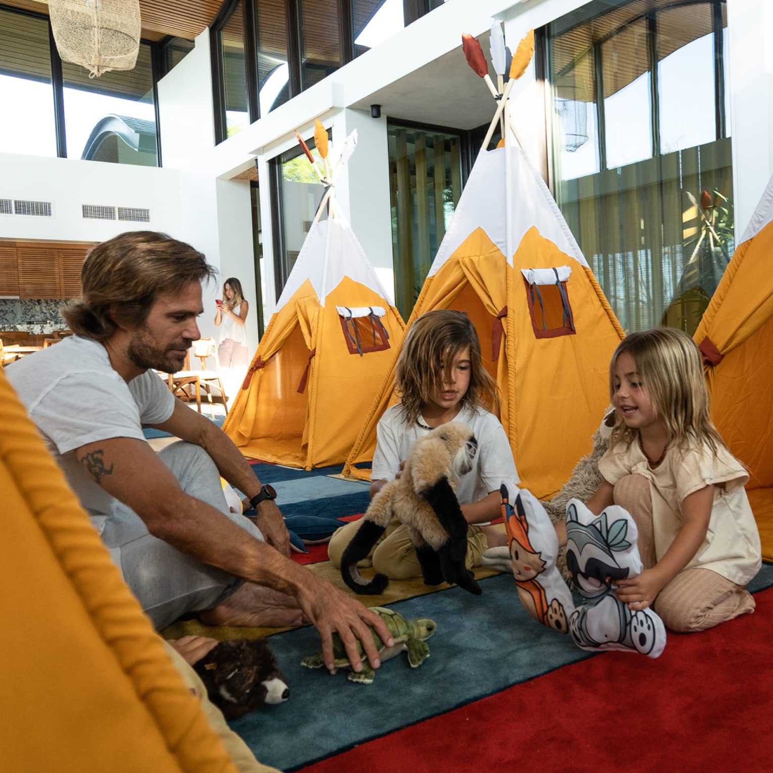 Two young kids and an adult play with stuffed animals near a row of orange indoor play tents