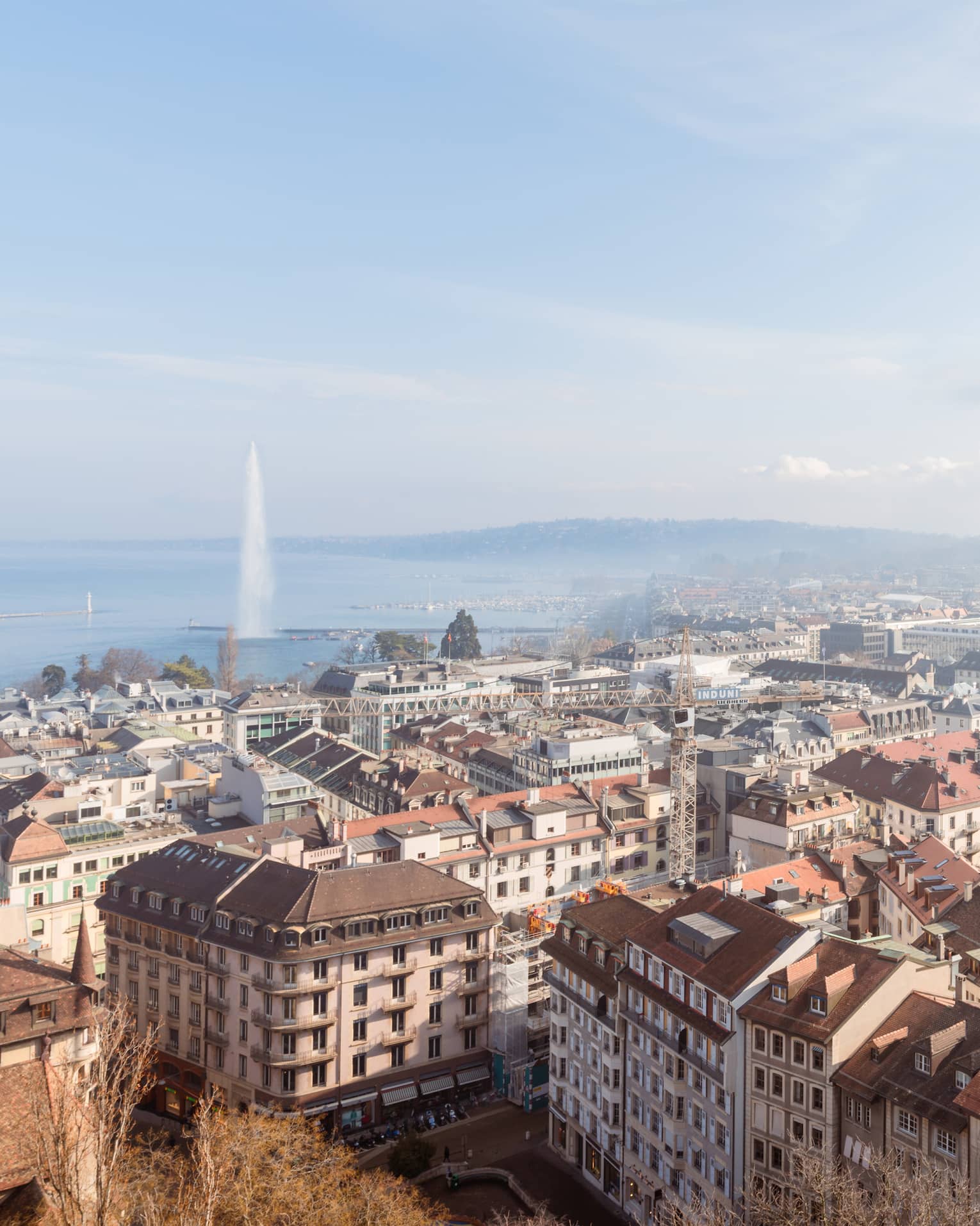 A cityscape with rooftops, buildings and streets, with a large fountain shooting water into the air over a lake in the background under a clear blue sky